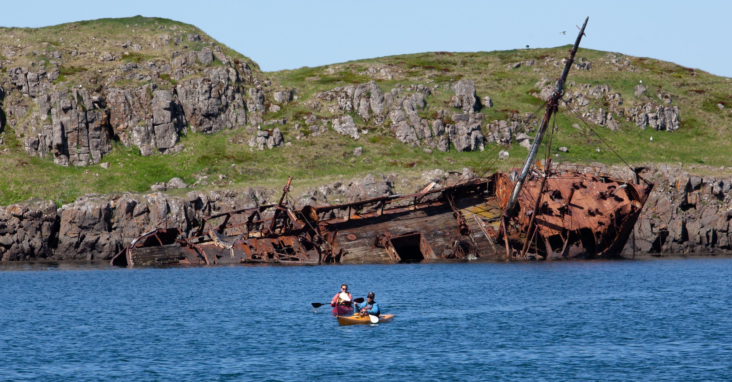 Two kayakers following a rocky West Iceland shore below a rusted shipwreck