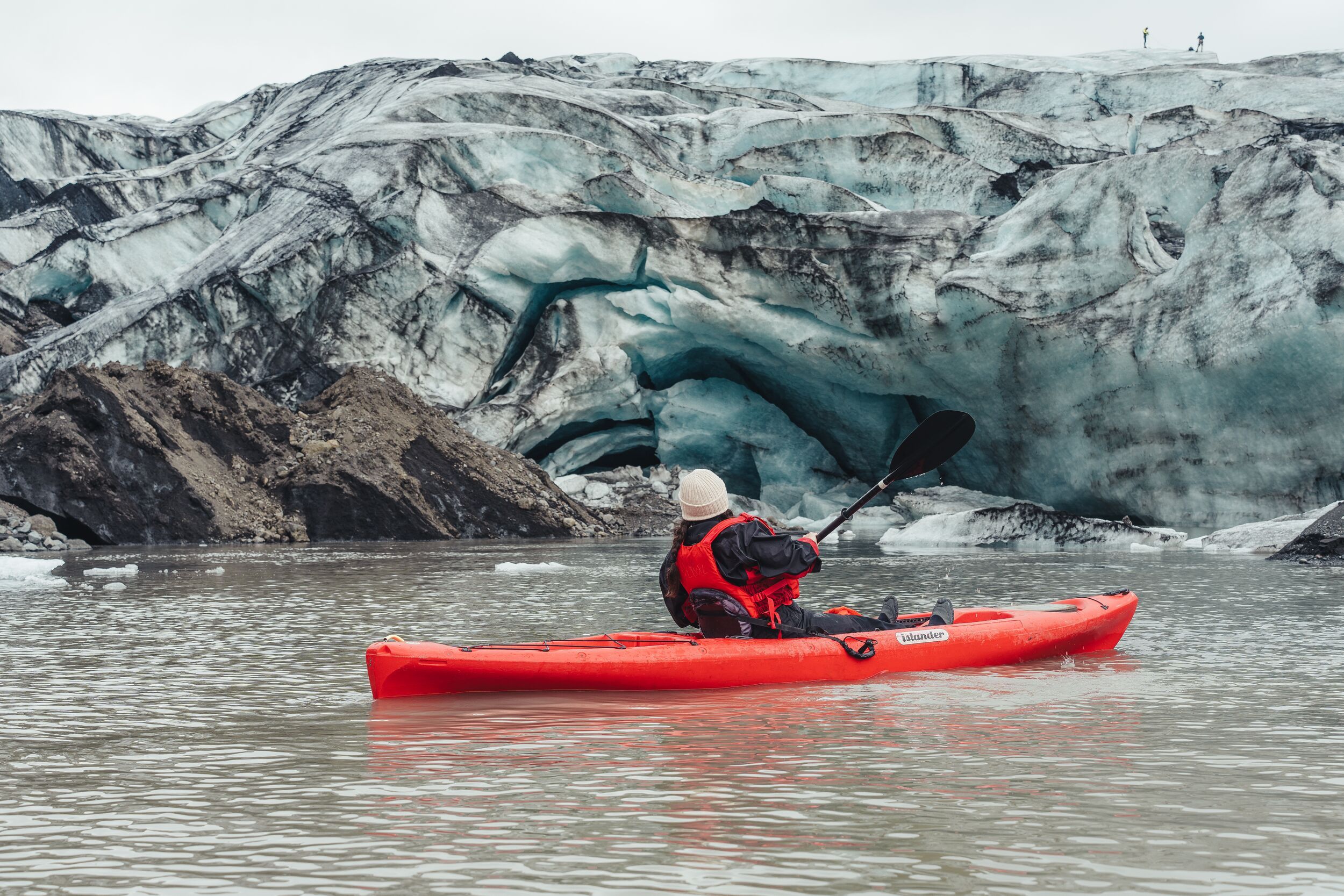A paddler in a red kayak facing folded blue and black glacier ice