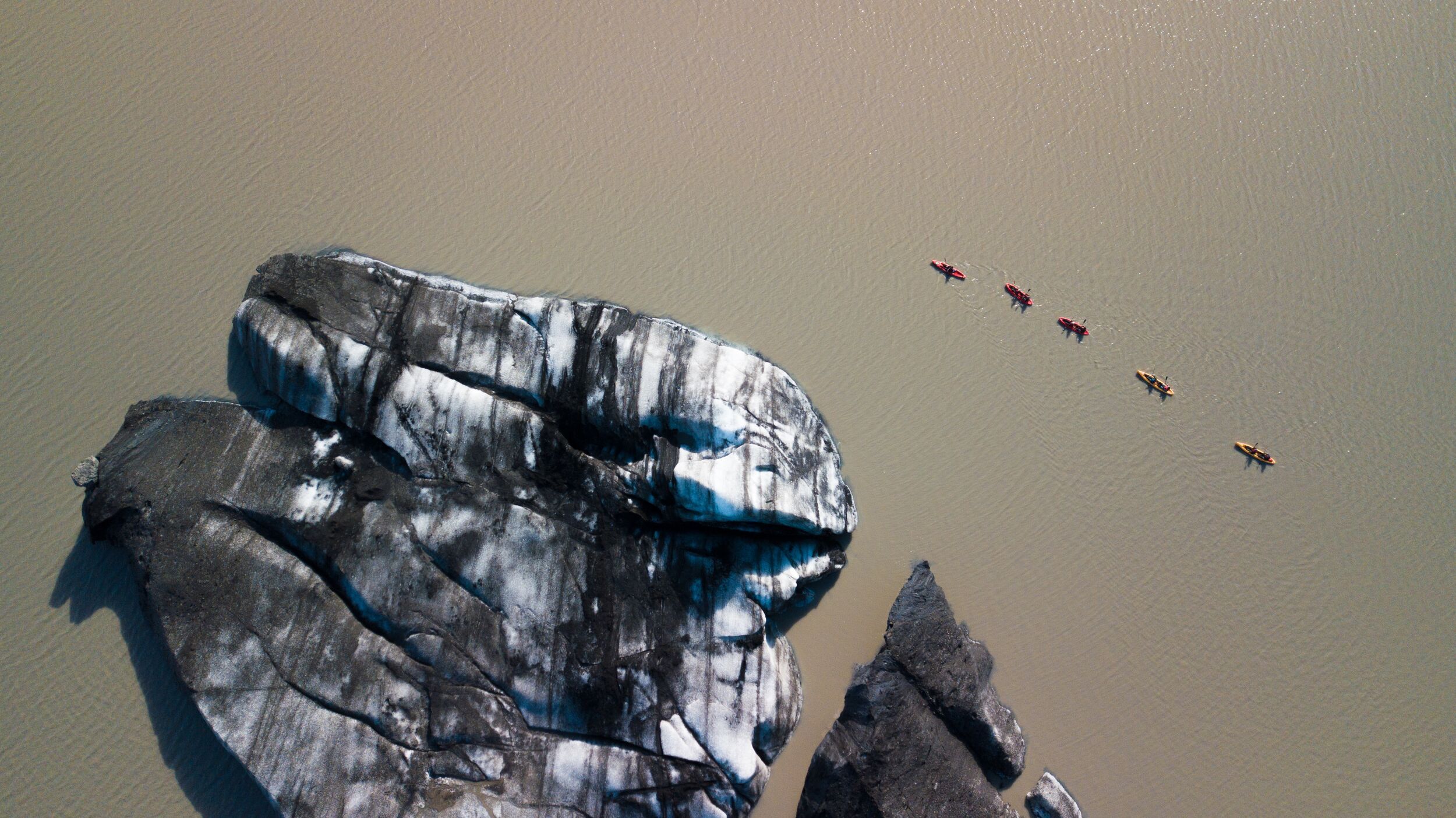 A line of small red and yellow kayaks passing a dark striped block of glacier ice