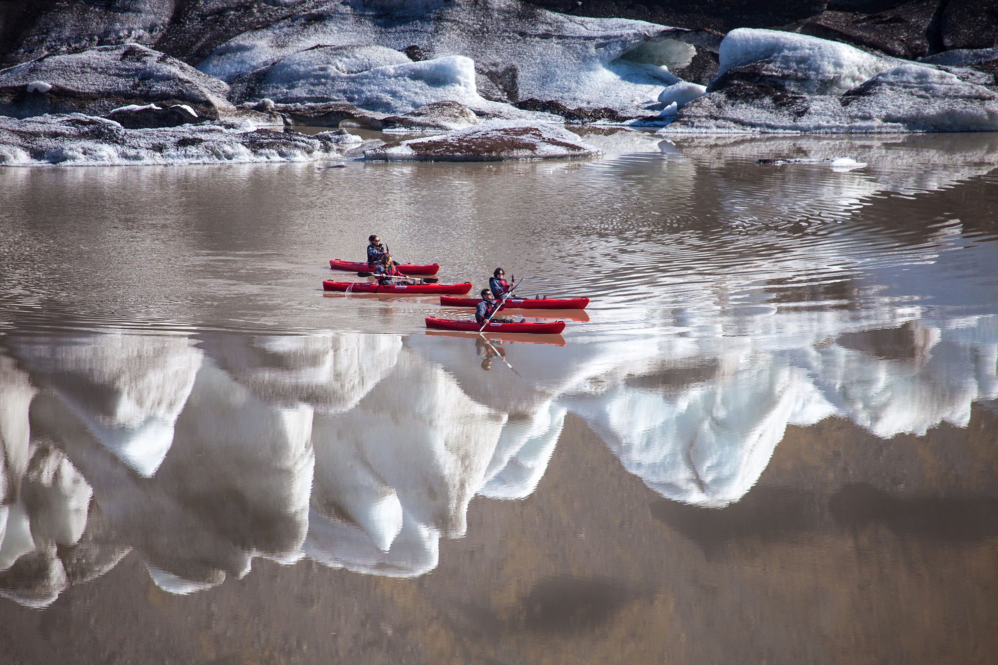 Five kayakers crossing reflective brown water below dark striped glacier ice