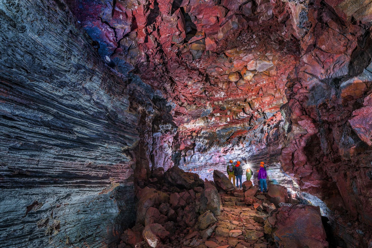 Helmeted visitors walking over rough lava rock inside the colorful walls of Raufarhólshellir
