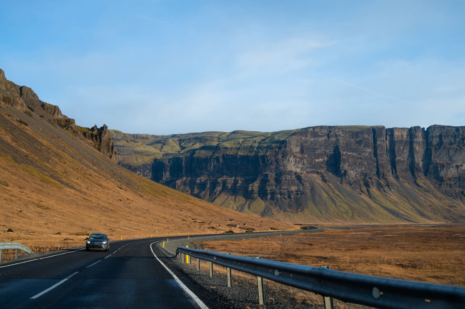Car traveling on a curving paved road below broad cliffs on Iceland's South Coast