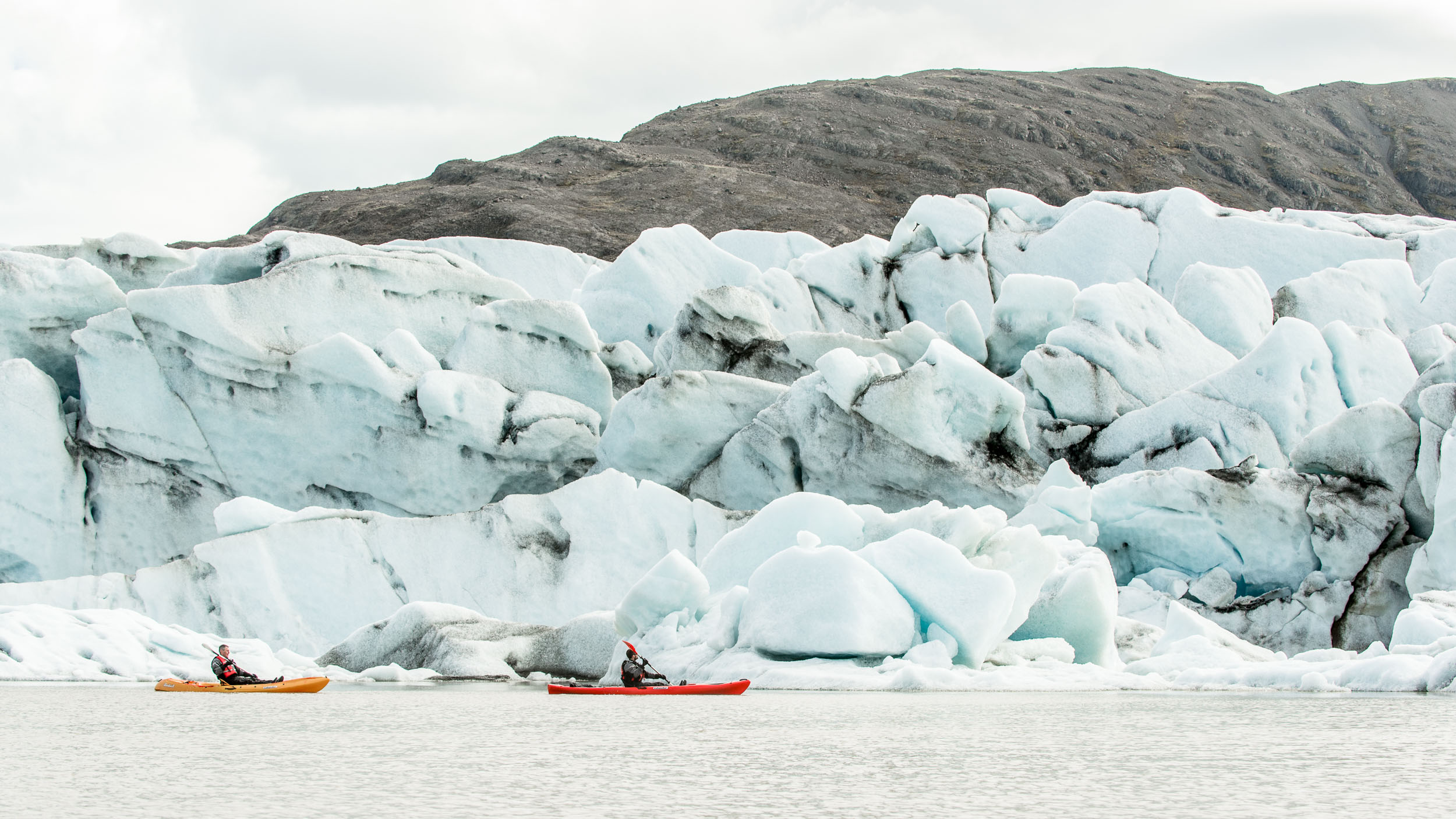 Two kayakers crossing a glacier lagoon below broken blue ice at Sólheimajökull