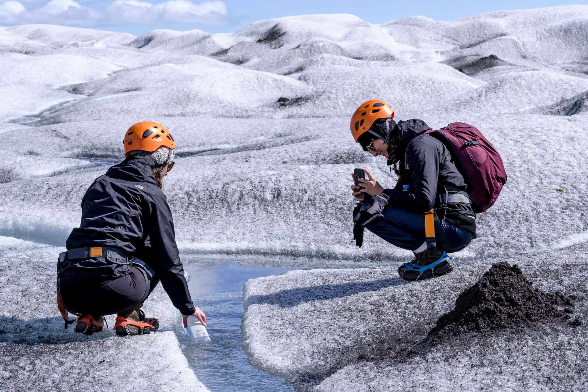 Two helmeted travelers in crampons pausing beside meltwater on an Icelandic glacier