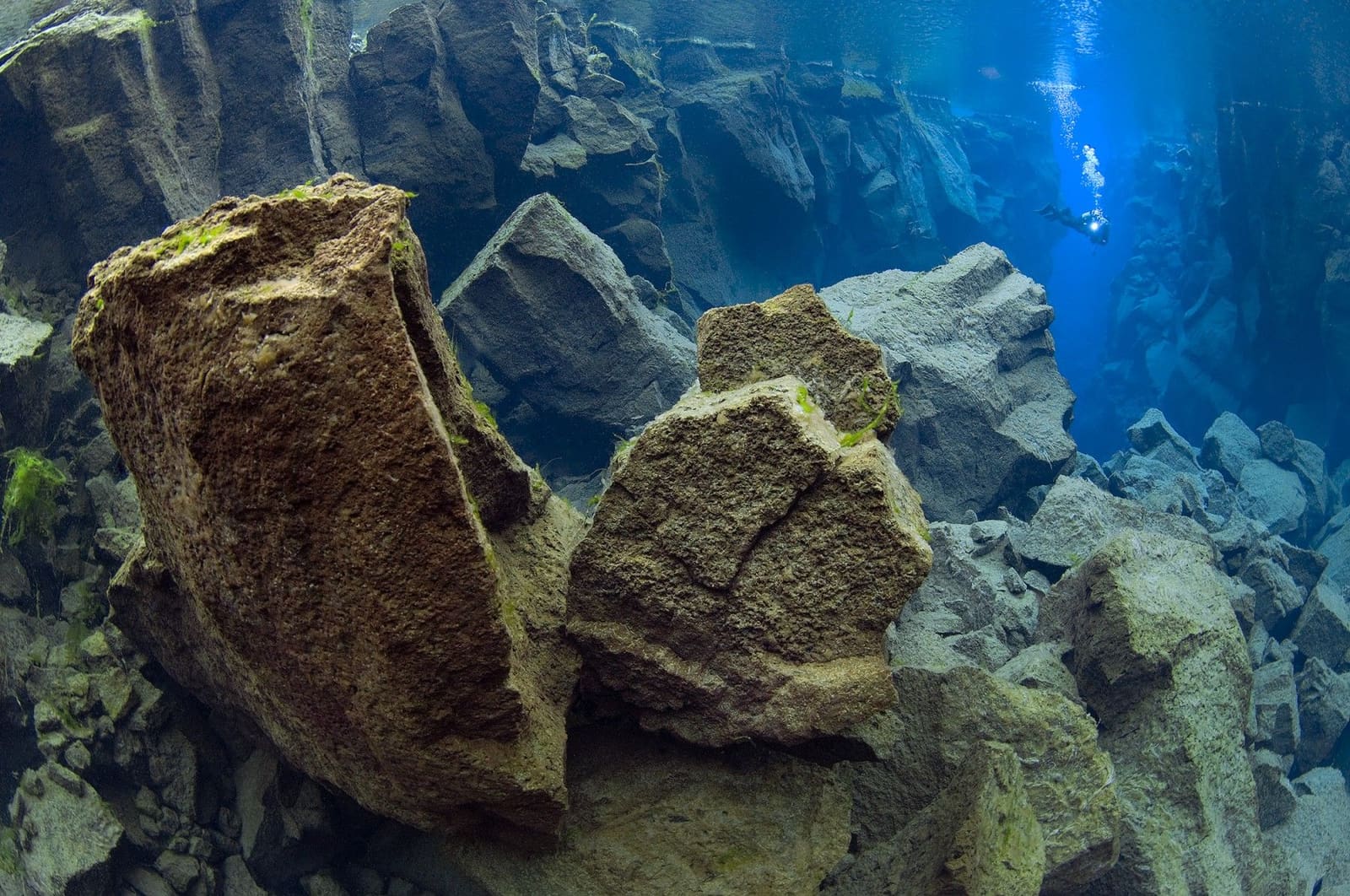 Diver leaving the Silfra Cathedral fissure through clear blue water and lava rock.