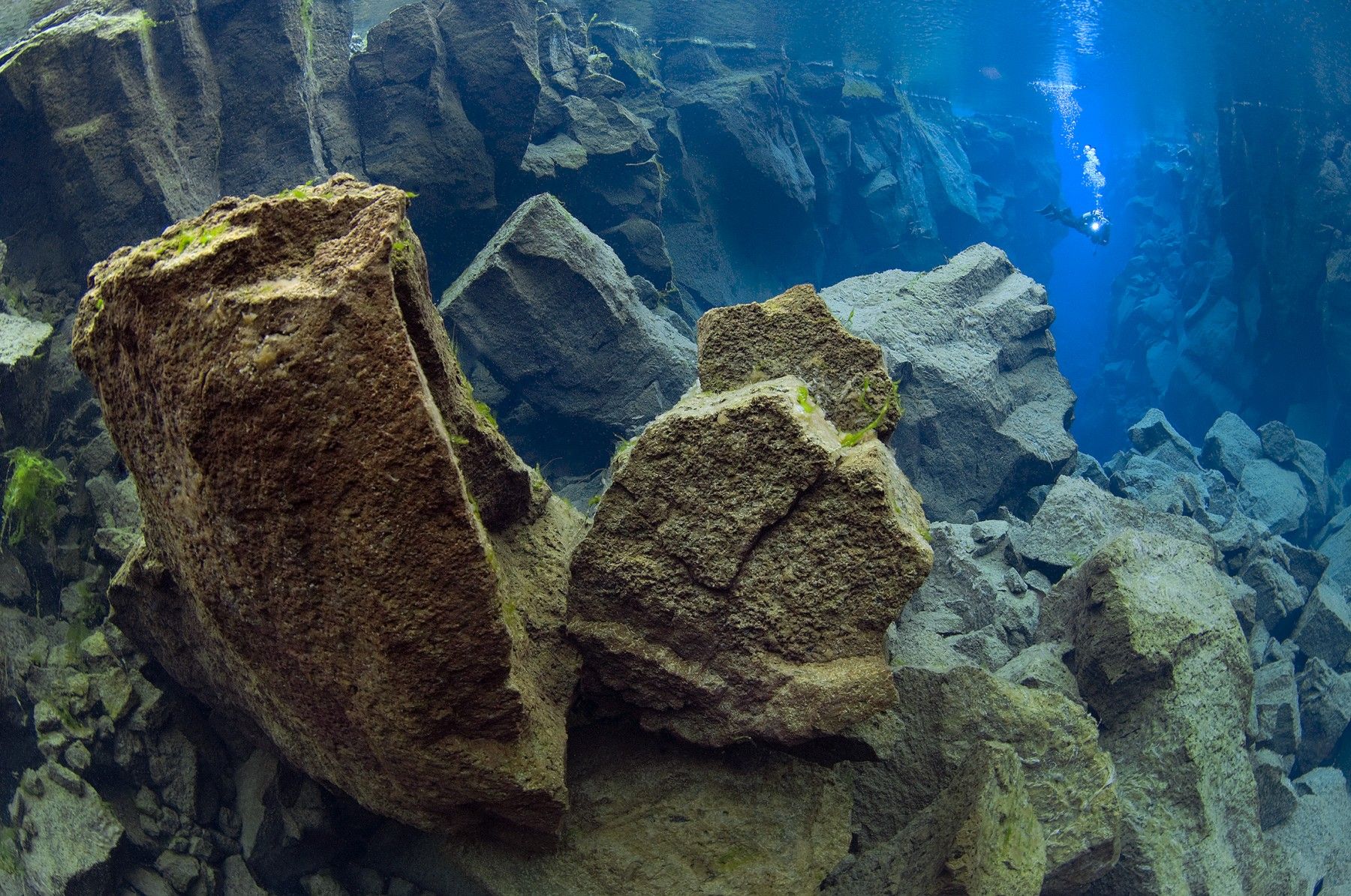 Diver leaving the Silfra Cathedral fissure through clear blue water and lava rock.