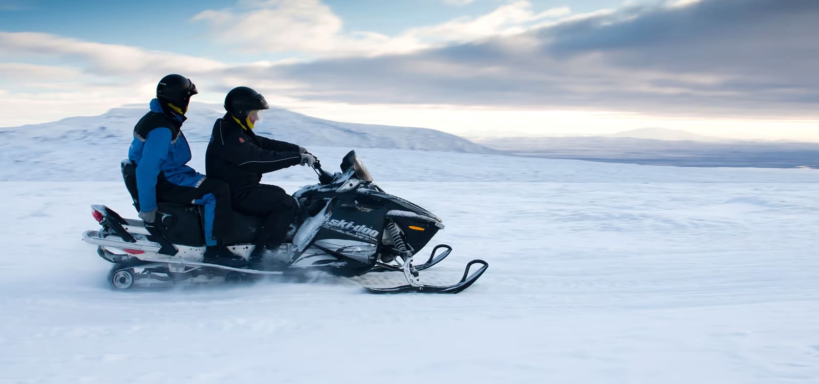Two helmeted travelers sharing a snowmobile across a broad Icelandic snowfield.