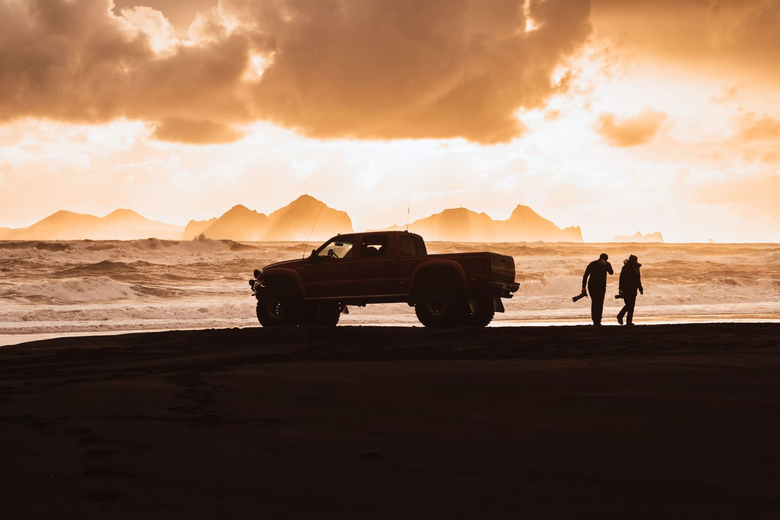 Modified pickup and two travelers silhouetted beside a black-sand coast at sunset