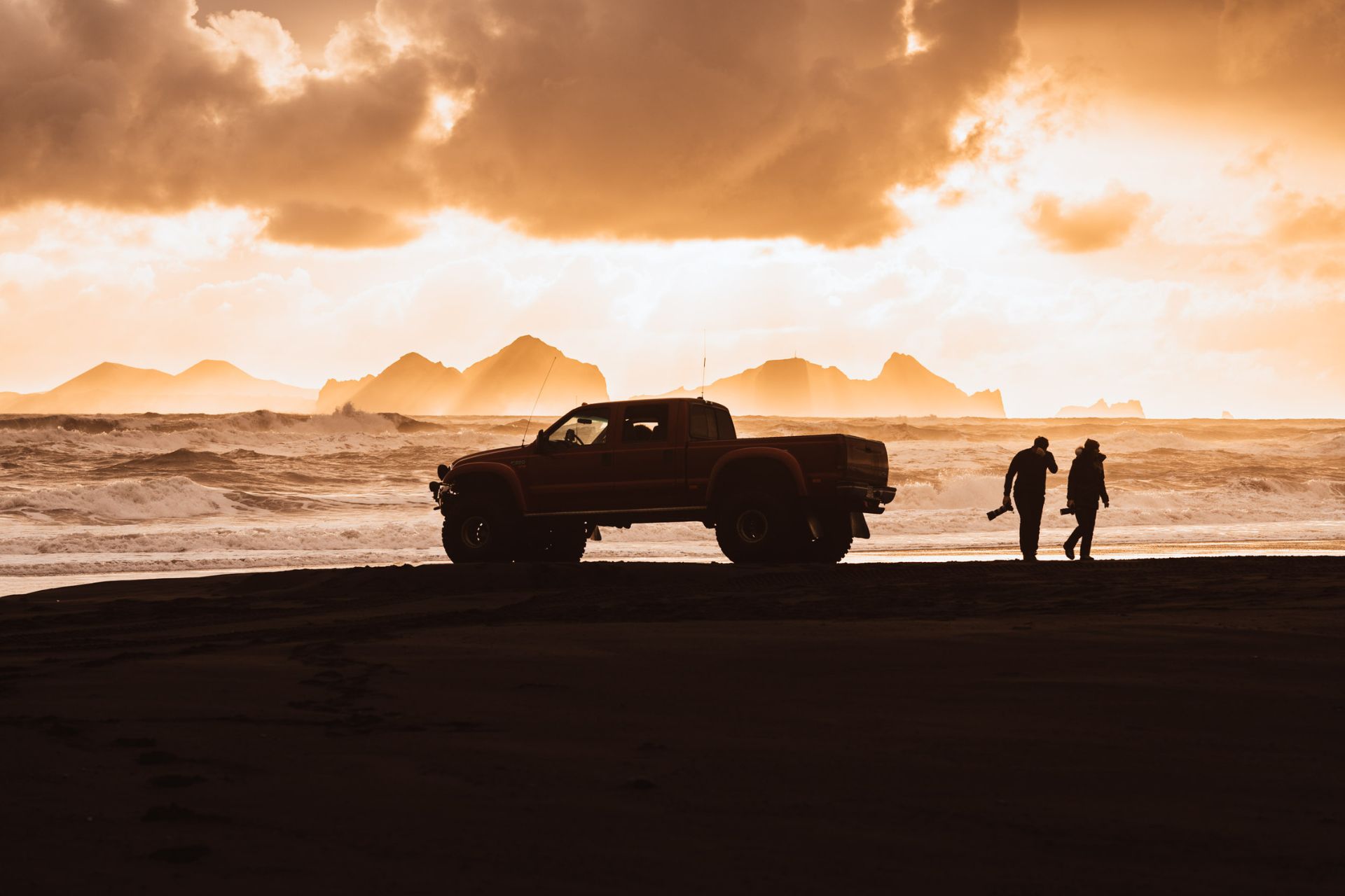 Modified pickup and two travelers silhouetted beside a black-sand coast at sunset