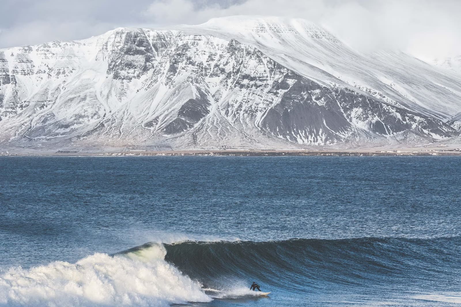 Surfer riding a dark wave below a snow-covered Icelandic mountain