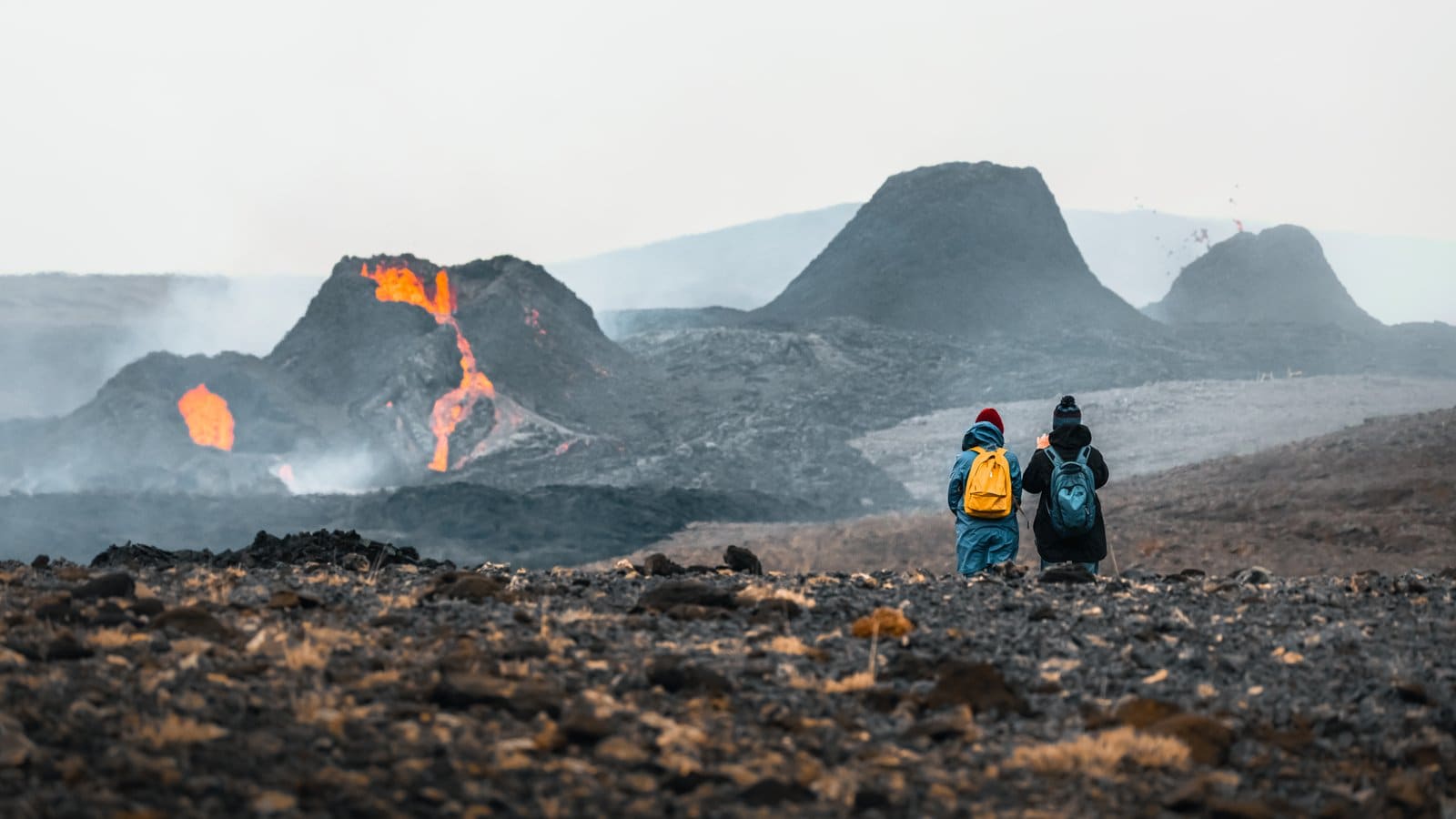 Two hikers watching the 2021 Geldingadalir eruption across dark volcanic ground in Iceland.