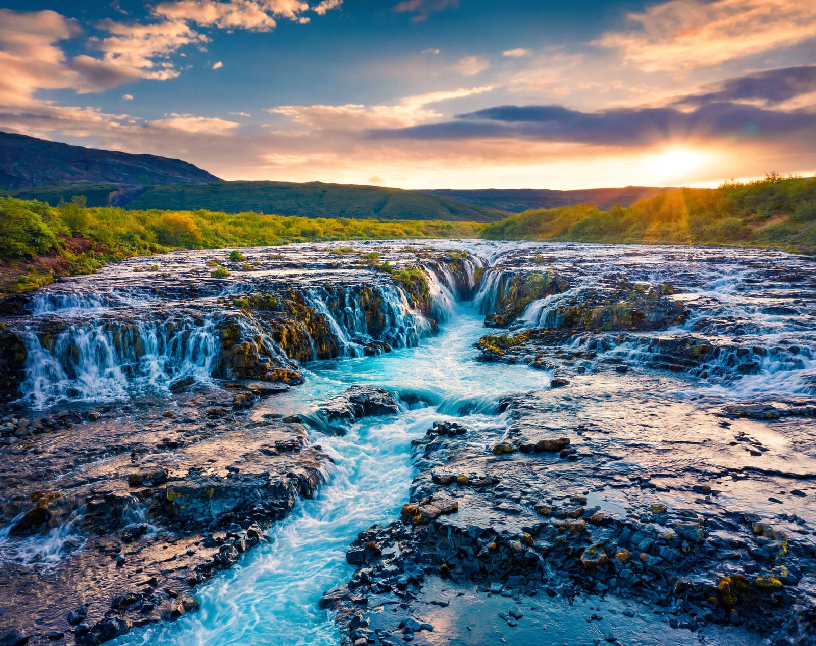 Brúarfoss Waterfall flowing through dark rock with vivid blue water at sunset.