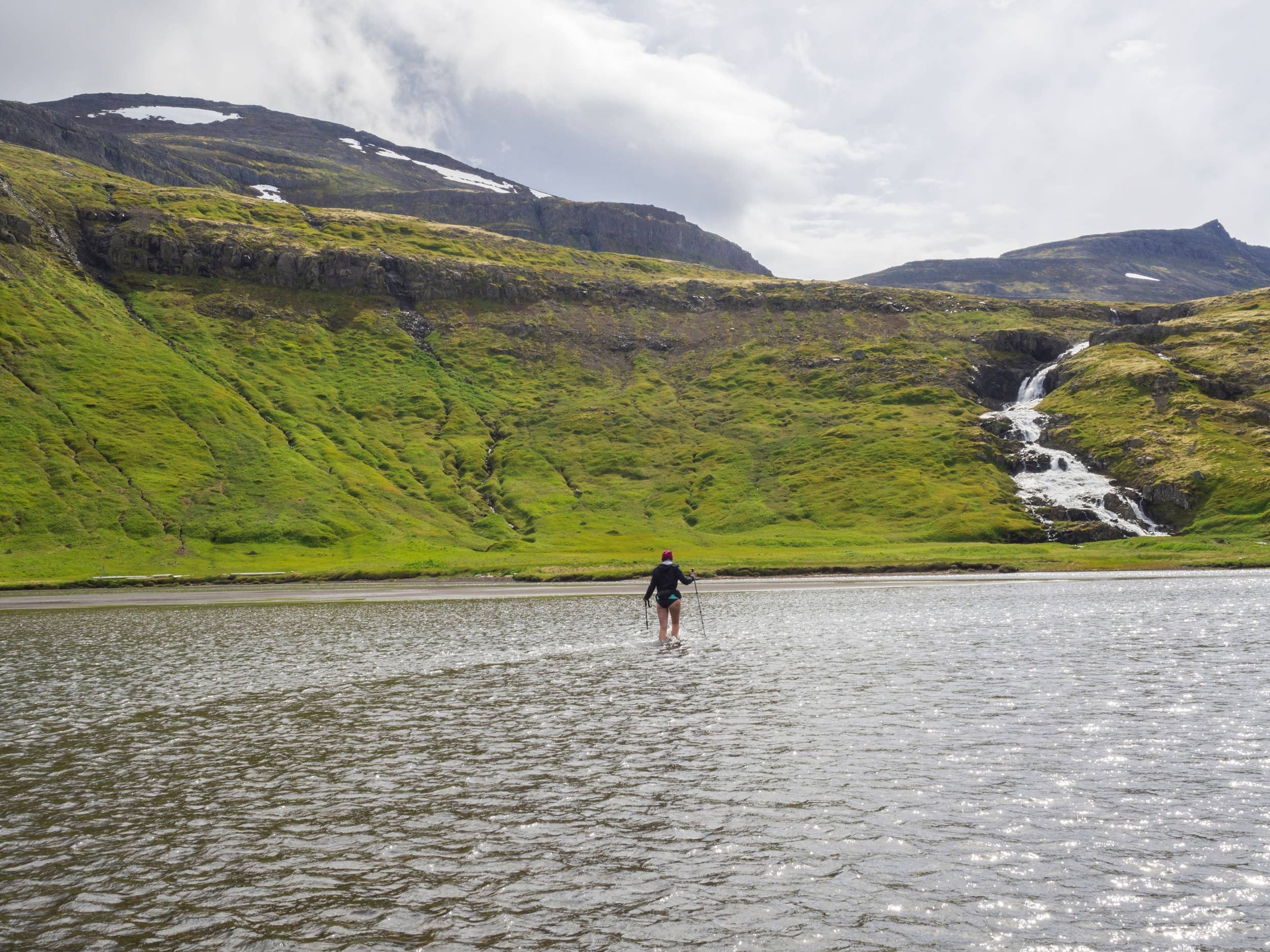 Hiker crossing a shallow river in Hornstrandir below green mountain slopes.