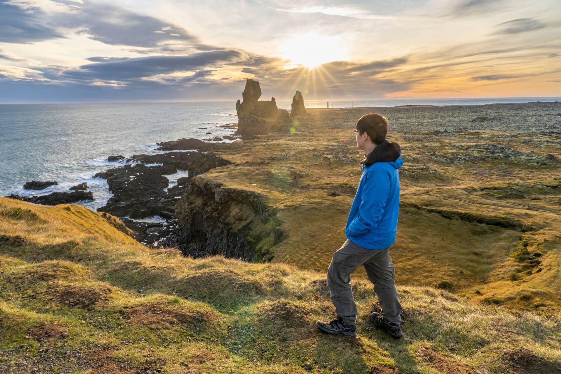 Traveler walking above sea cliffs at sunset in Snæfellsjökull National Park with Atlantic coastline below.