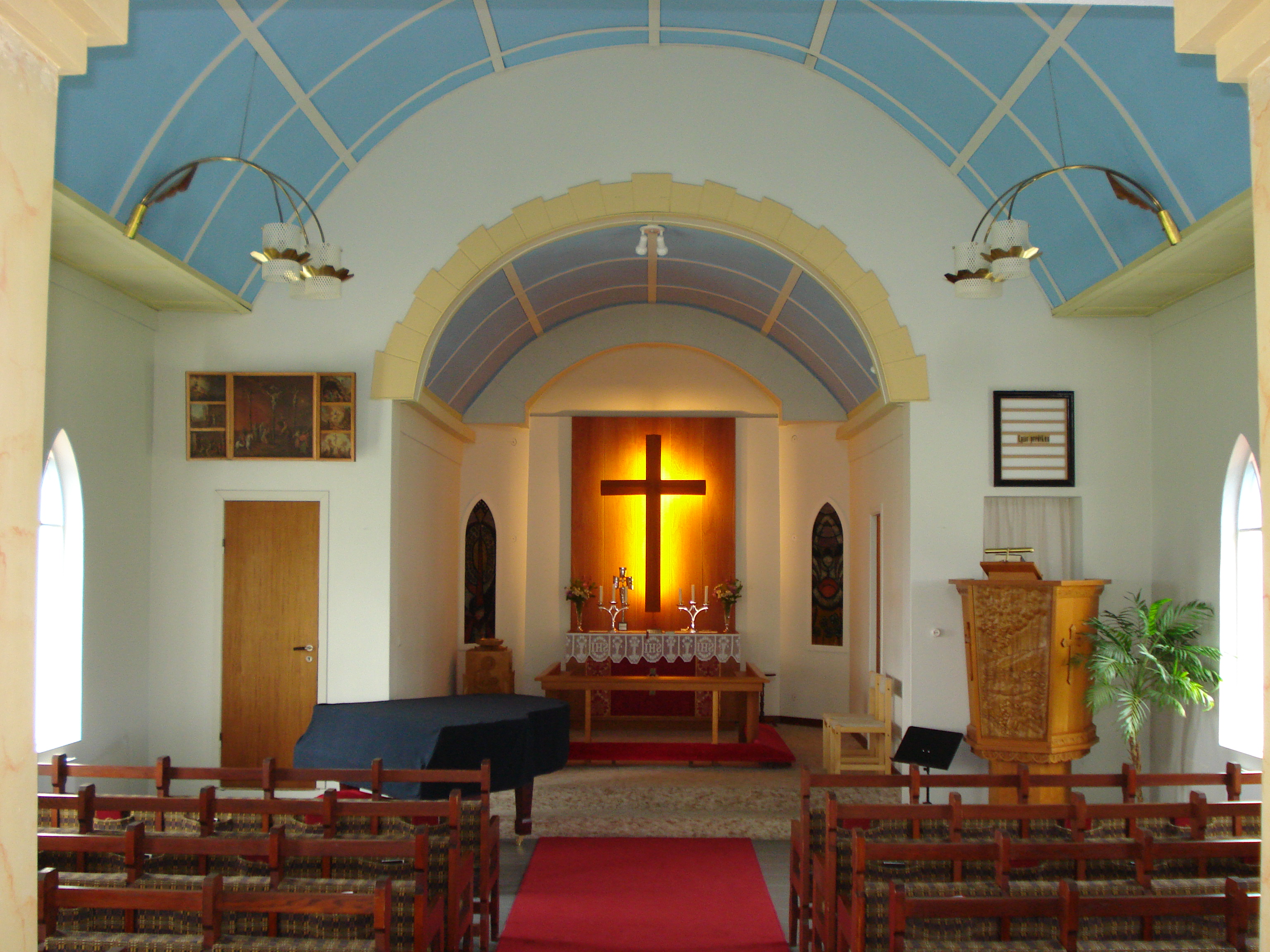 Interior of Reykjahlíðarkirkja with pale blue ceiling panels and a glowing cross above the altar.