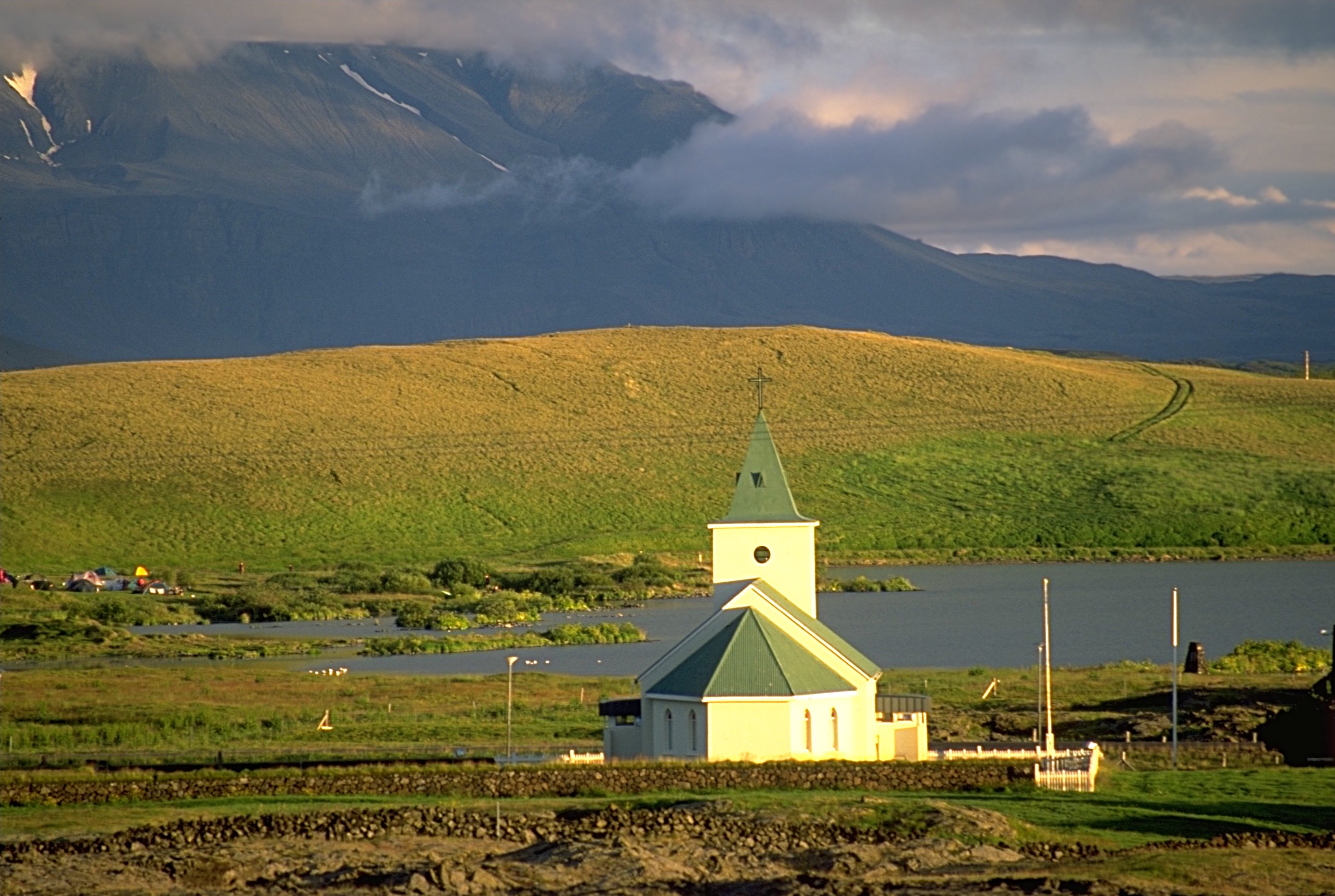 Reykjahlíðarkirkja above the lakeshore with green hills and a dark mountain ridge behind.