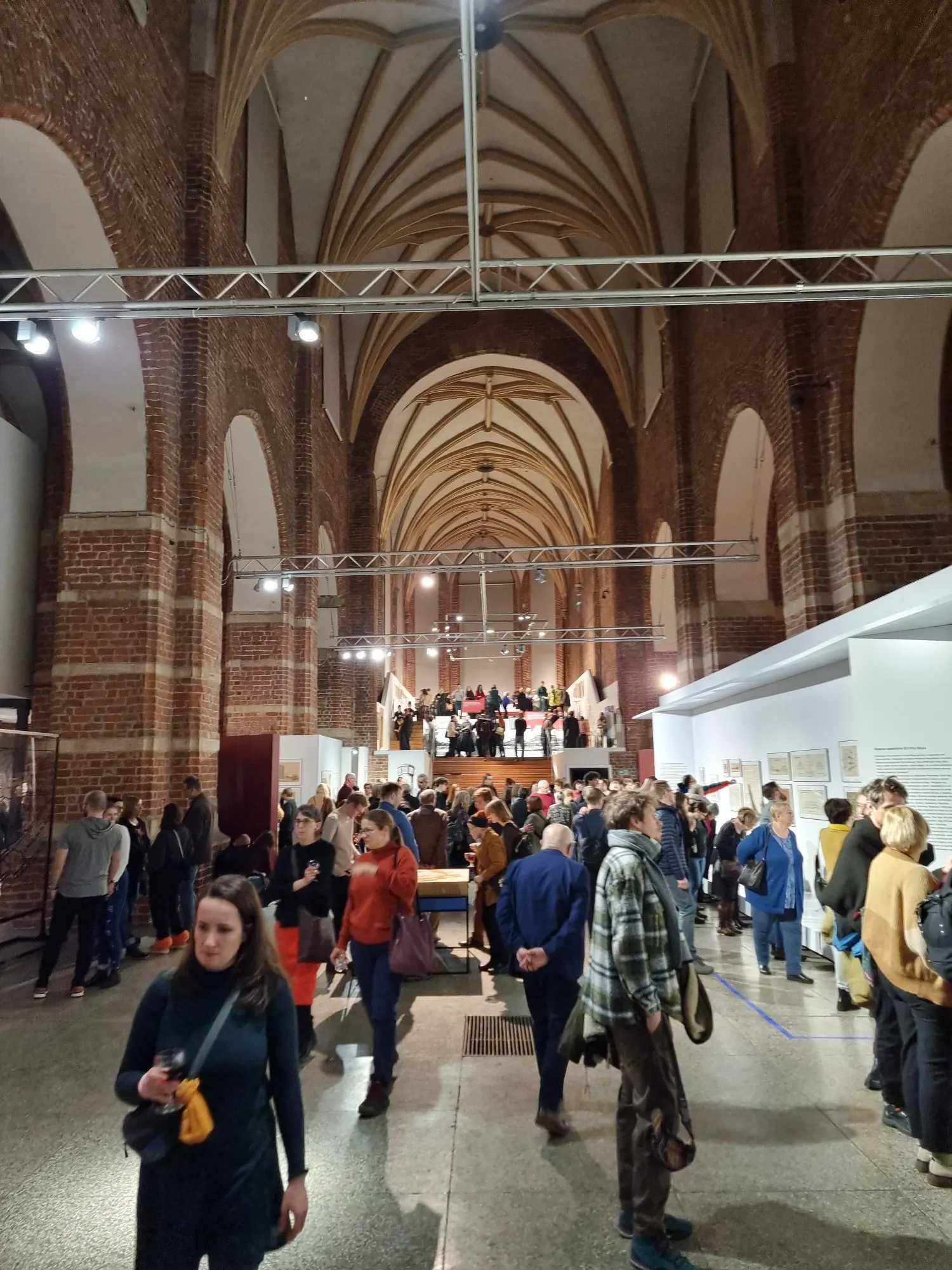 A crowd of people at an art exhibition inside a historic brick building with high, Gothic vaulted ceilings.