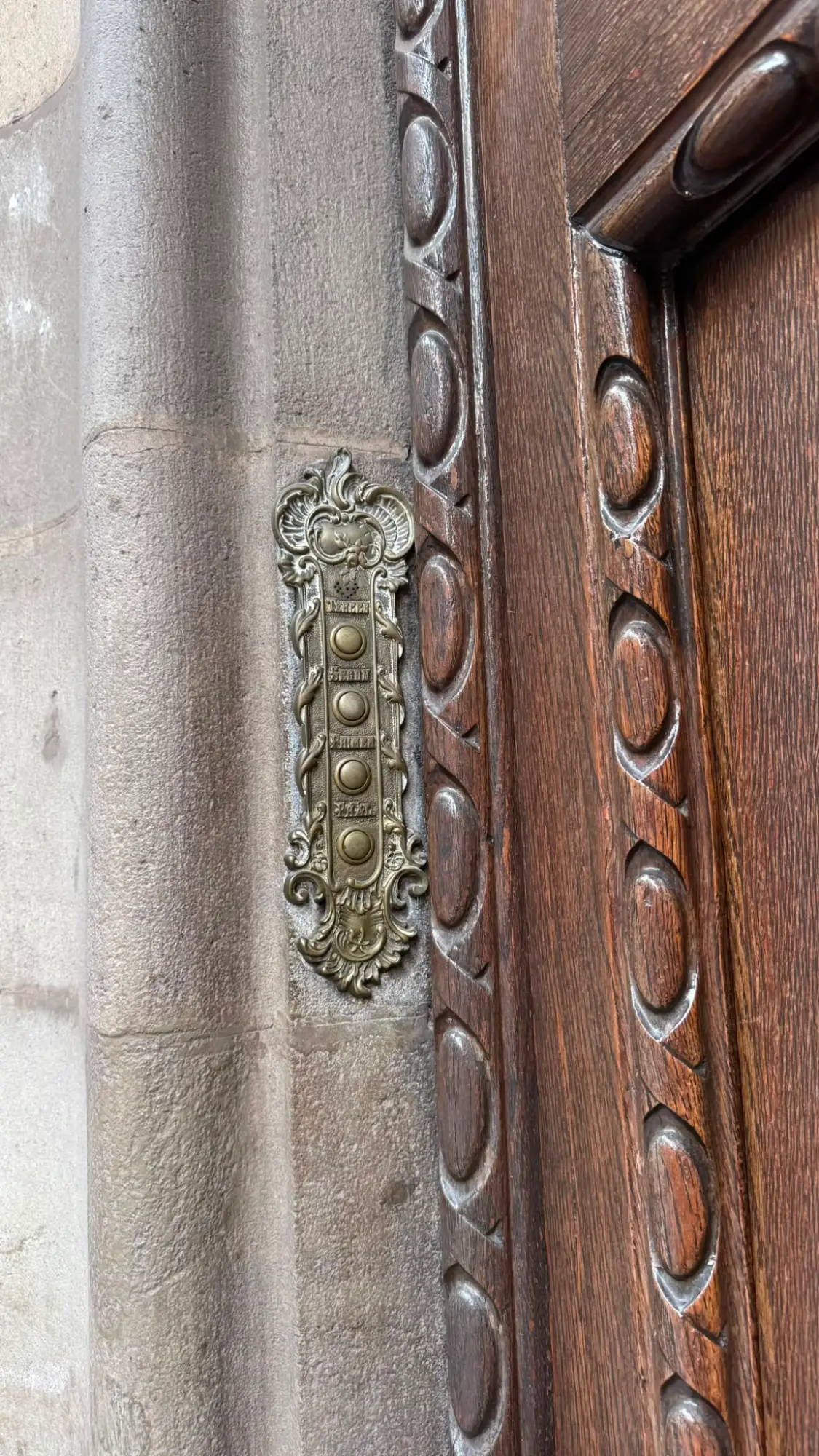 An ornate, vertical brass doorbell panel with four buttons on a stone wall next to a dark wooden door.
