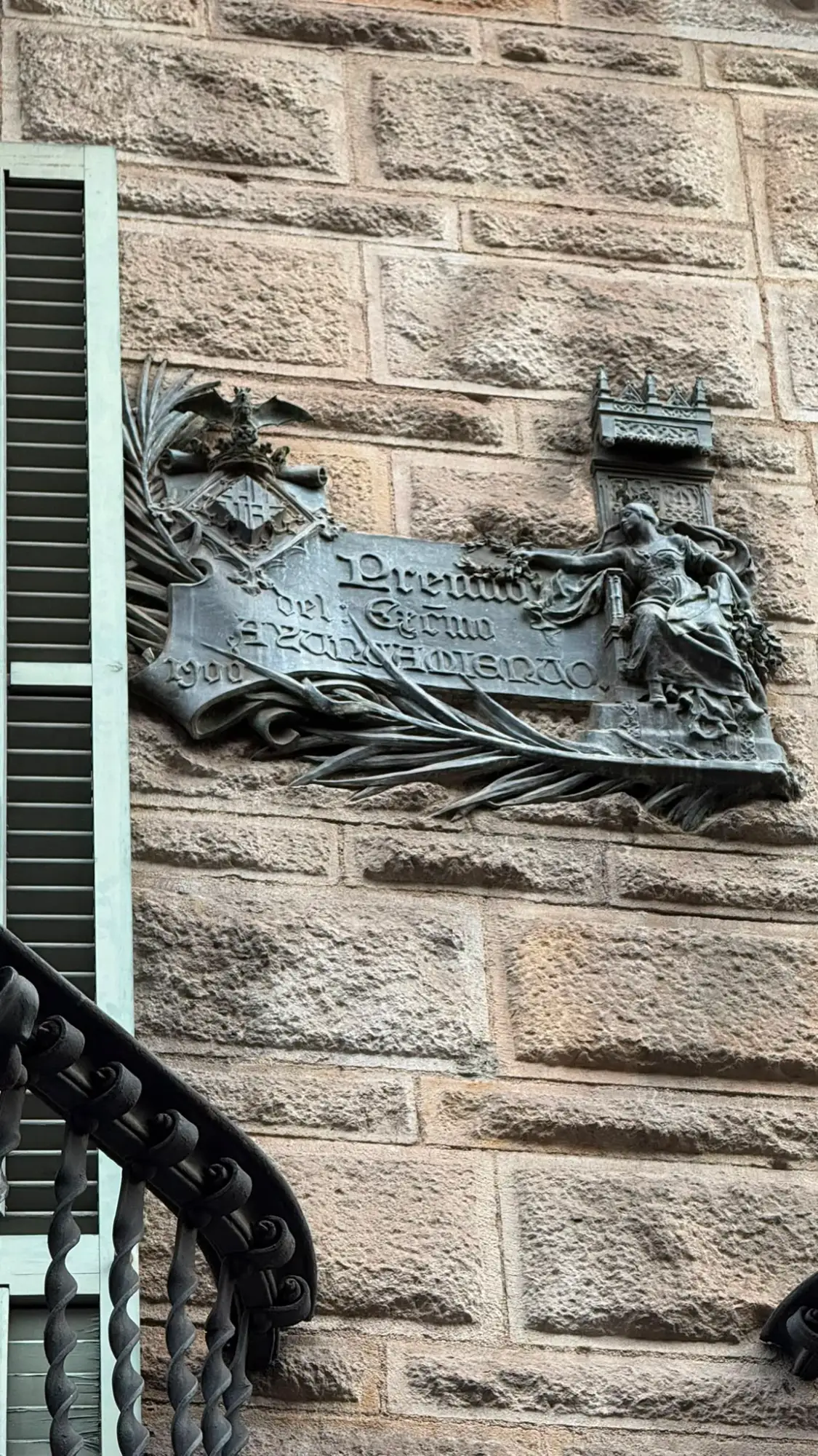 An ornate Art Nouveau-style metal plaque from the year 1900 on a stone building wall in Barcelona.