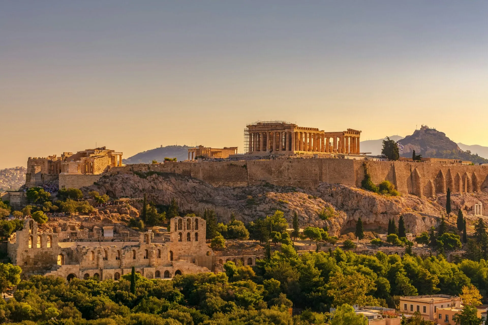 Die Akropolis von Athen bei Sonnenuntergang mit dem Parthenon, dem Erechtheion und dem Odeon des Herodes Atticus, umgeben von Bäumen.