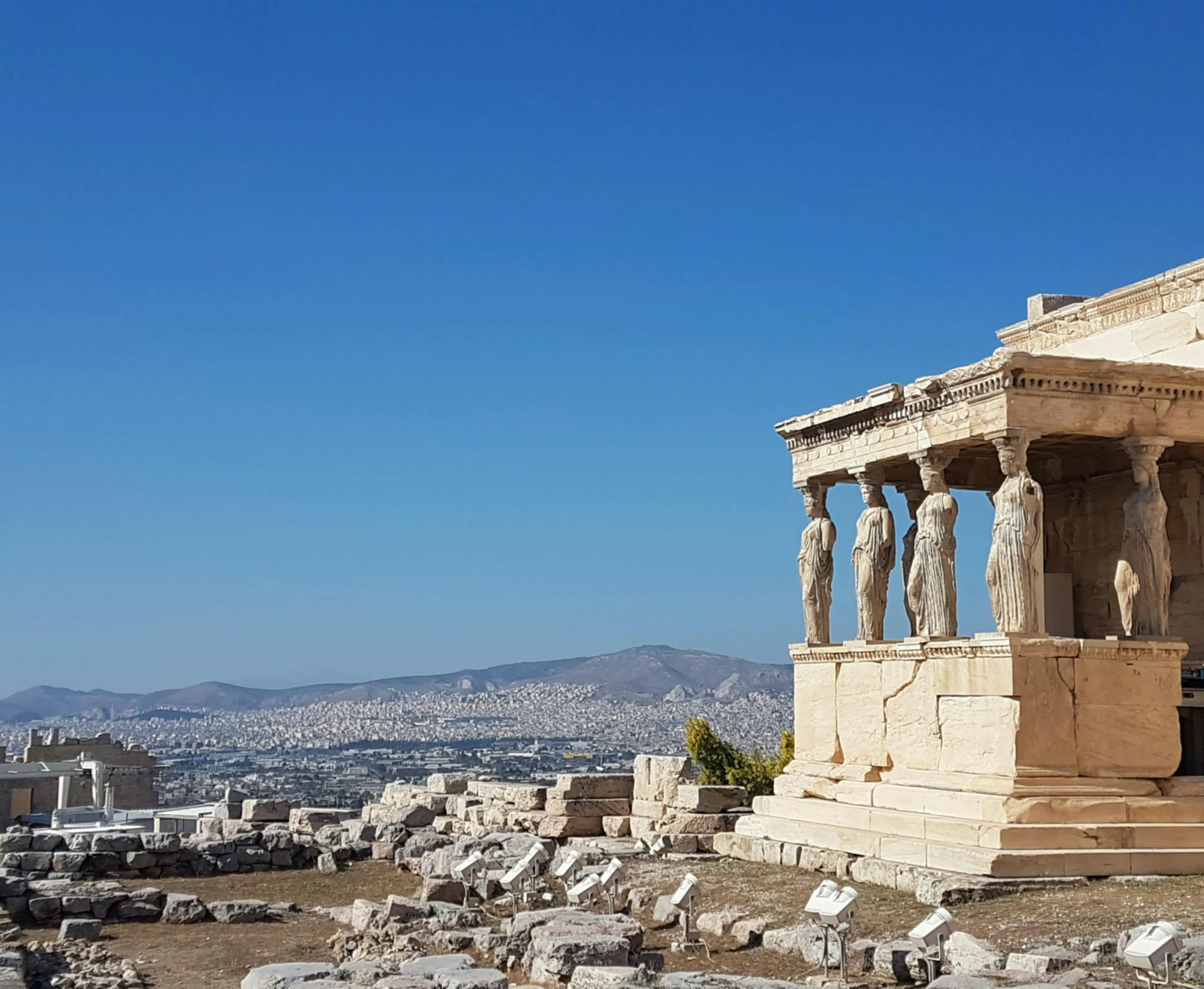 The Porch of the Caryatids at the Erechtheion on the Acropolis, with Athens visible in the background under a clear blue sky.