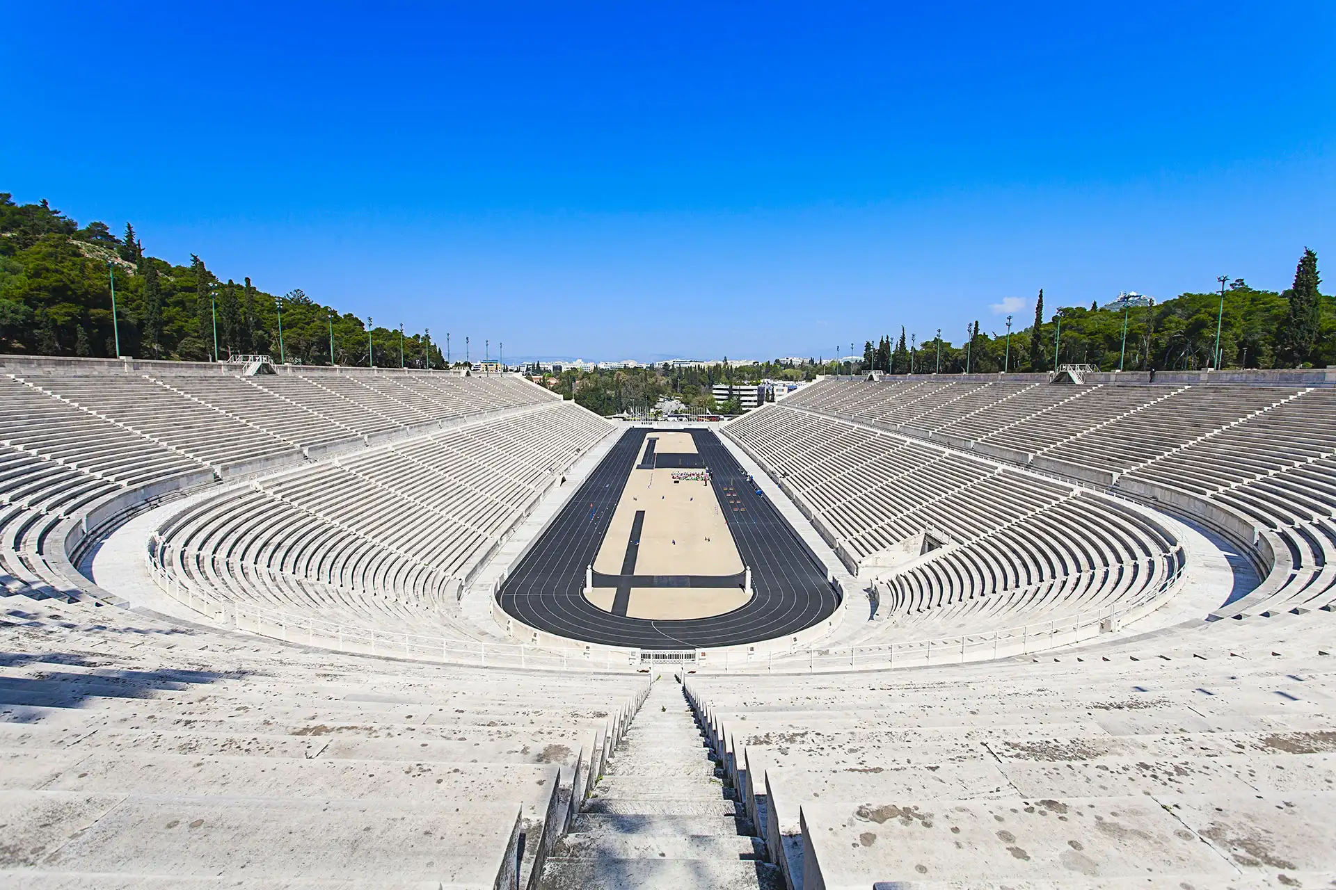 The Panathenaic Stadium in Athens, a U-shaped structure of white marble seats, viewed from above on a clear, sunny day.