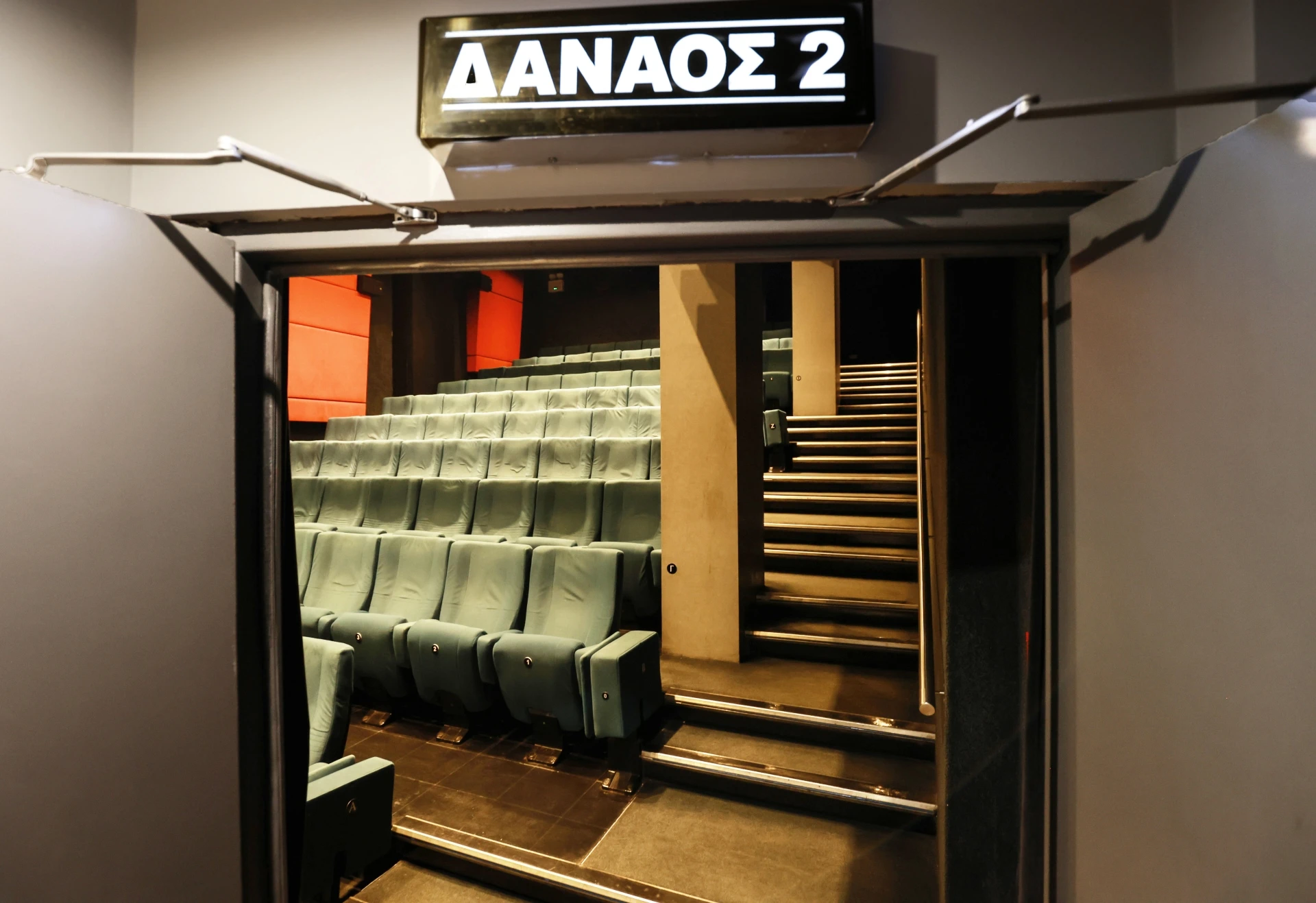 View through an open doorway into an empty screening room with rows of green cinema seats and stairs under a Danaos 2 sign.