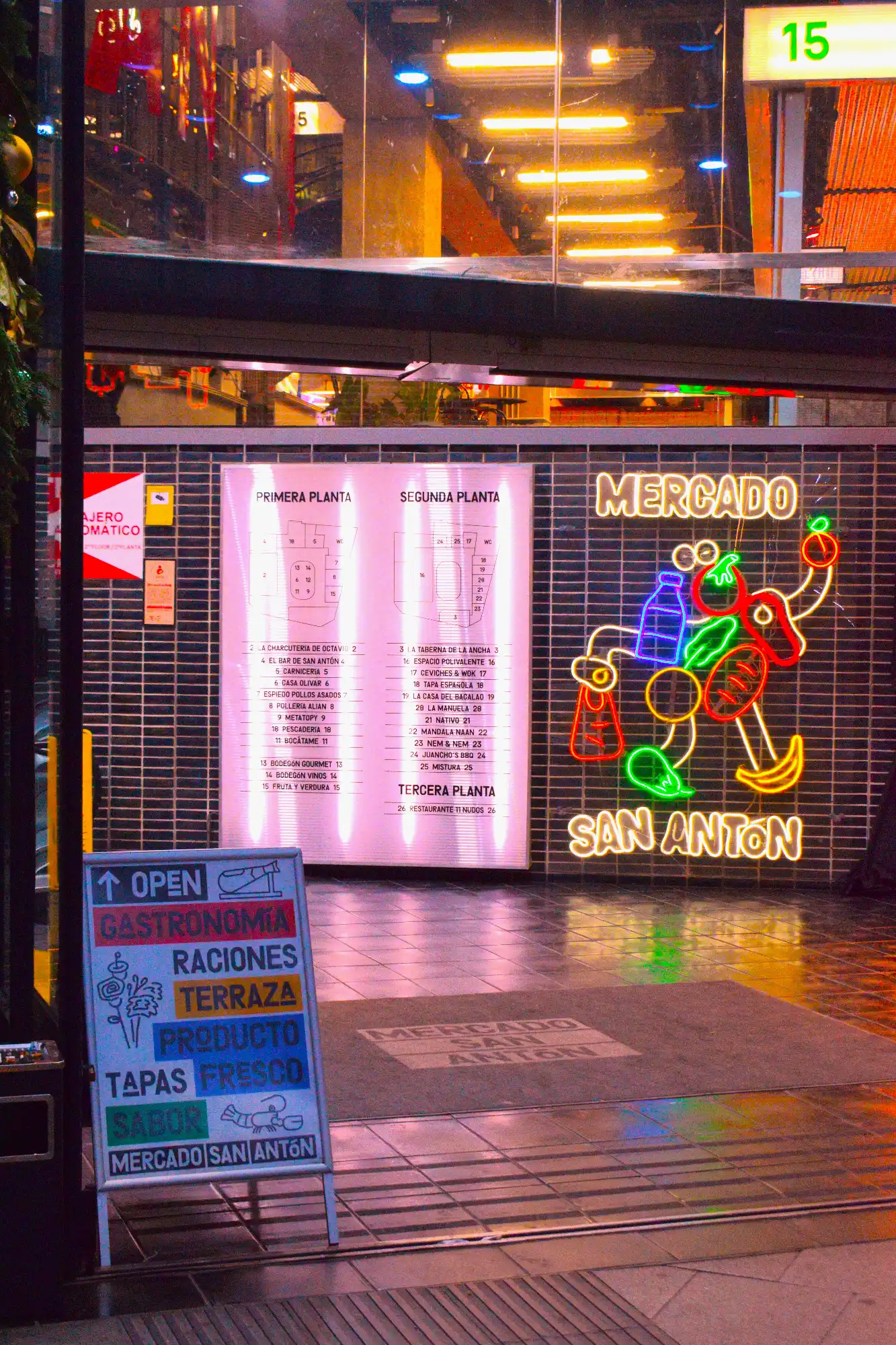 The illuminated entrance to Mercado San Antón at night, with a colourful neon sign and a directory of food stalls.