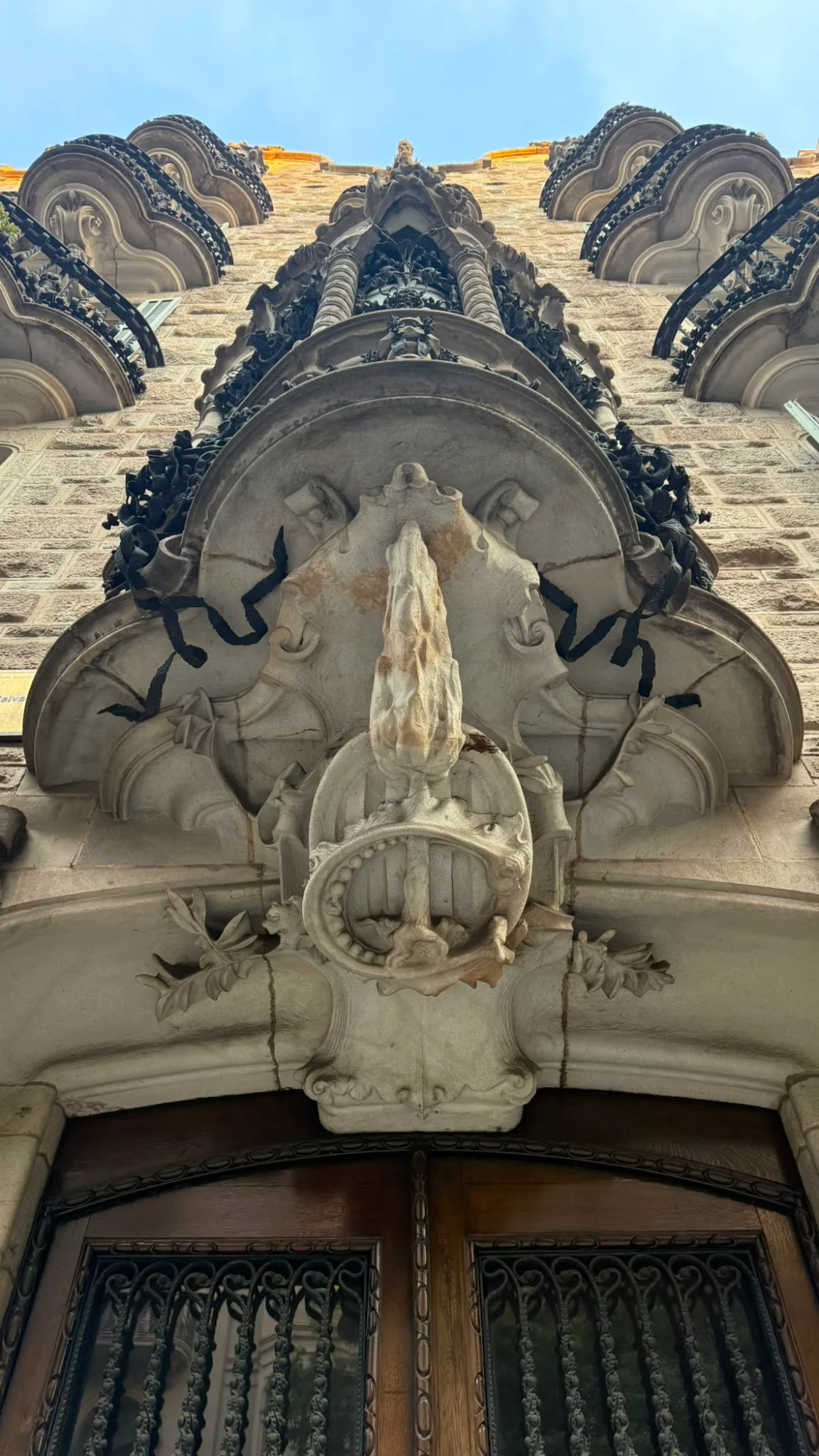 A low-angle view of a stone building facade with an elaborate sculpture over a doorway and wrought-iron balconies.