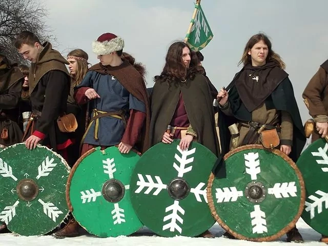 Group of historical reenactors in medieval costumes holding green round shields with white symbols in a snowy landscape.