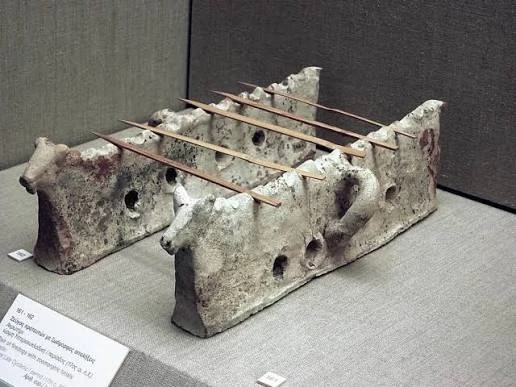 Two ancient Greek ceramic souvlaki trays with animal heads and holes for embers, displayed in a museum.