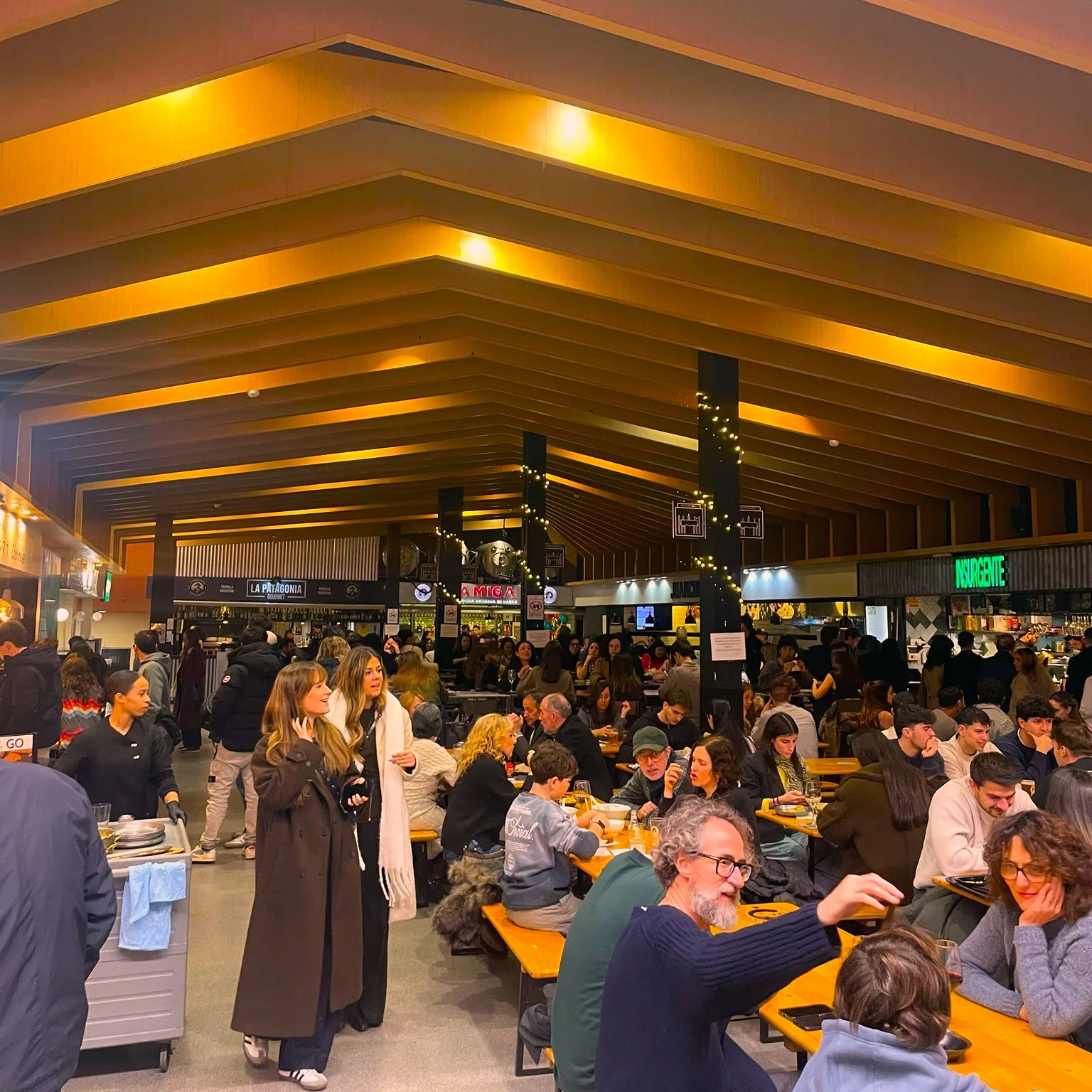 A busy, modern food hall with a high wooden ceiling, filled with people eating and socializing at long communal tables.