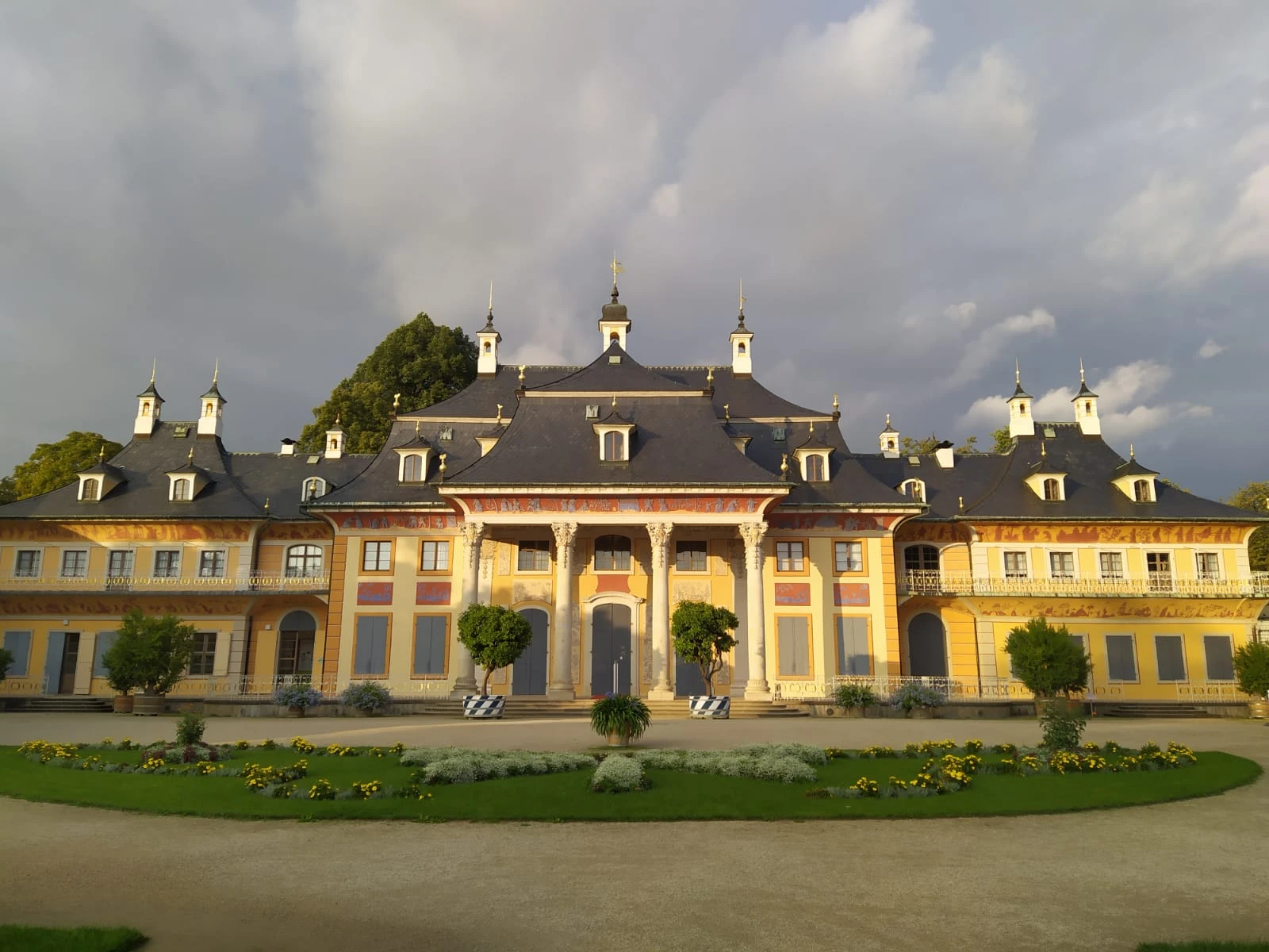 The Riverside Palace at Pillnitz, Dresden, under a dramatic, cloudy sky, with manicured gardens in the foreground.