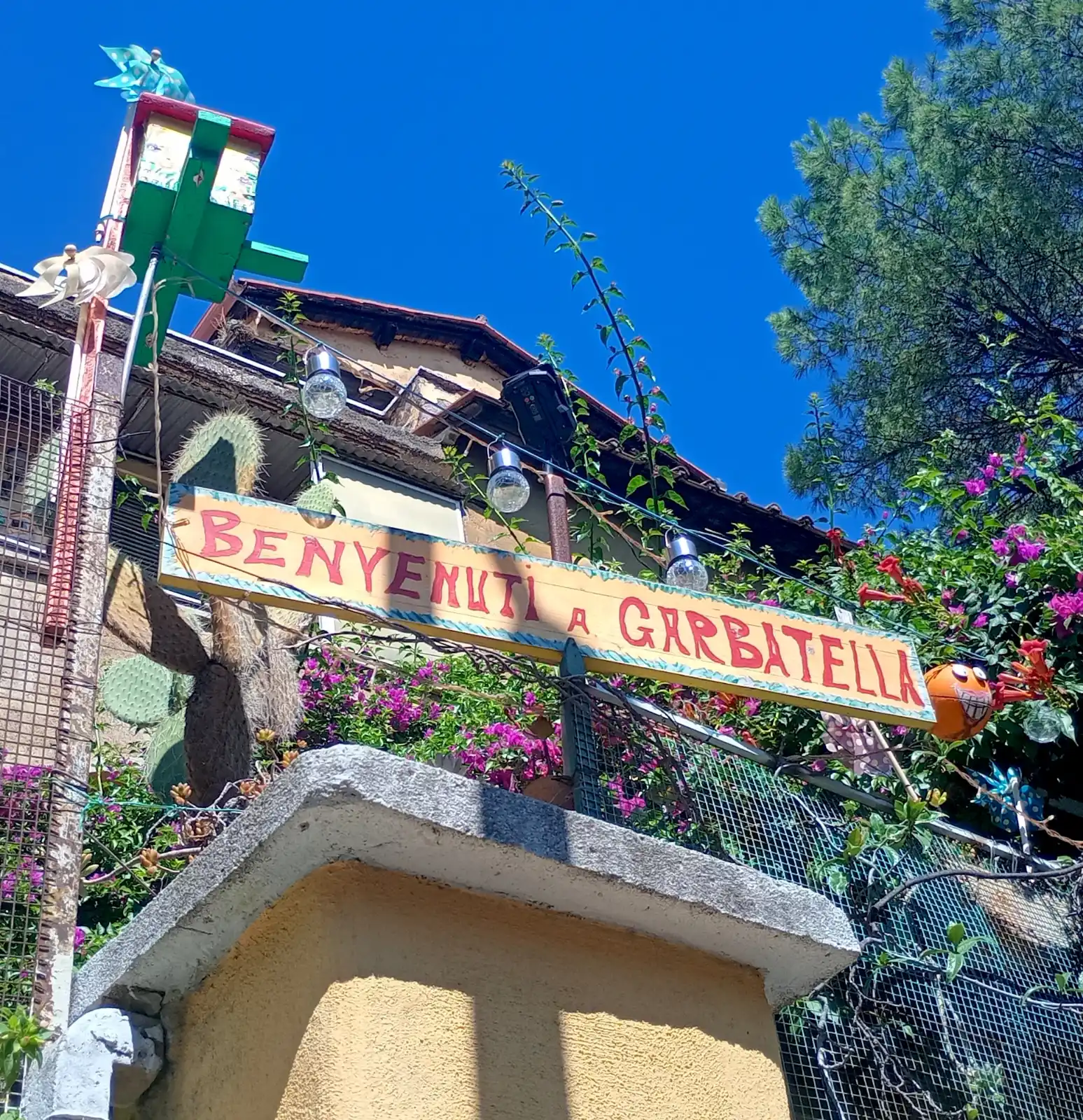 A colourful, hand-painted "Benvenuti a Garbatella" sign surrounded by flowers and plants against a blue sky.