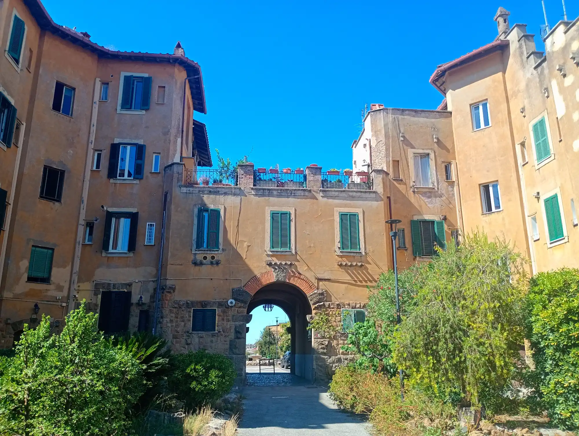 A stone archway connects ochre-colored residential buildings with green shutters under a clear blue sky in Rome.