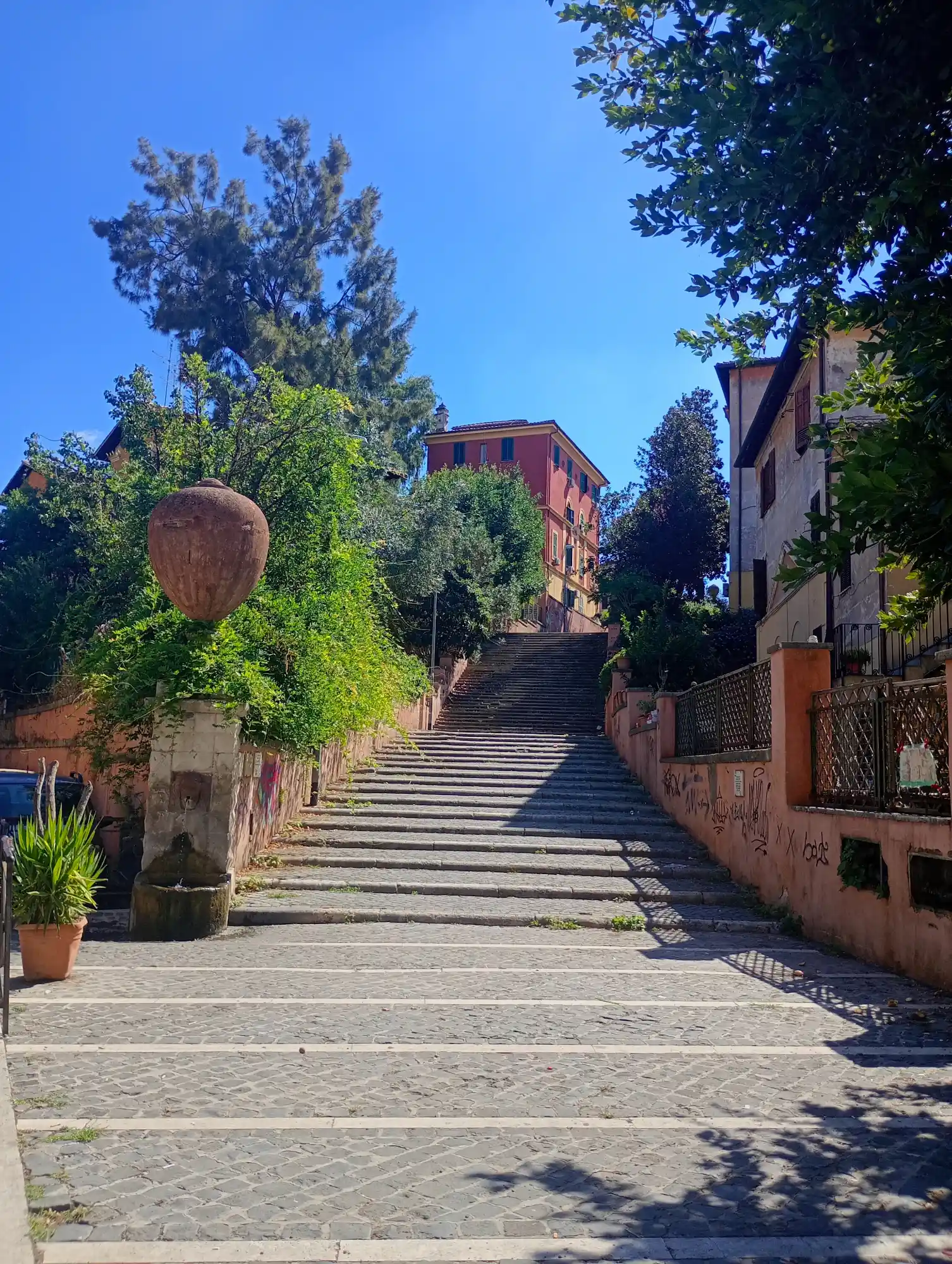 A wide stone staircase ascends a hill in Rome, flanked by buildings and lush greenery under a blue sky.