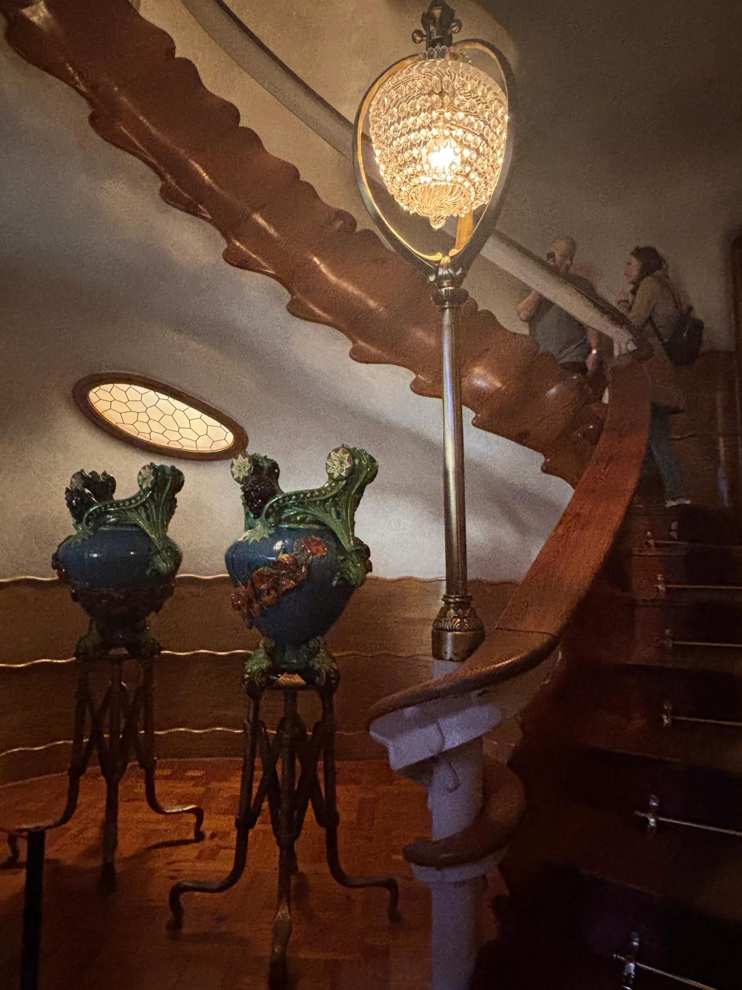 Curving wooden staircase in Casa Batlló with a crystal lamp, two large blue vases, and an oval window.