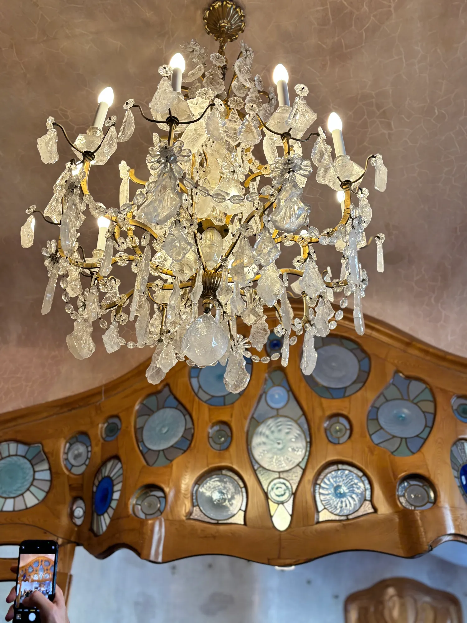 An ornate crystal chandelier hangs above a curved wooden frame with round stained-glass windows inside Casa Batlló.