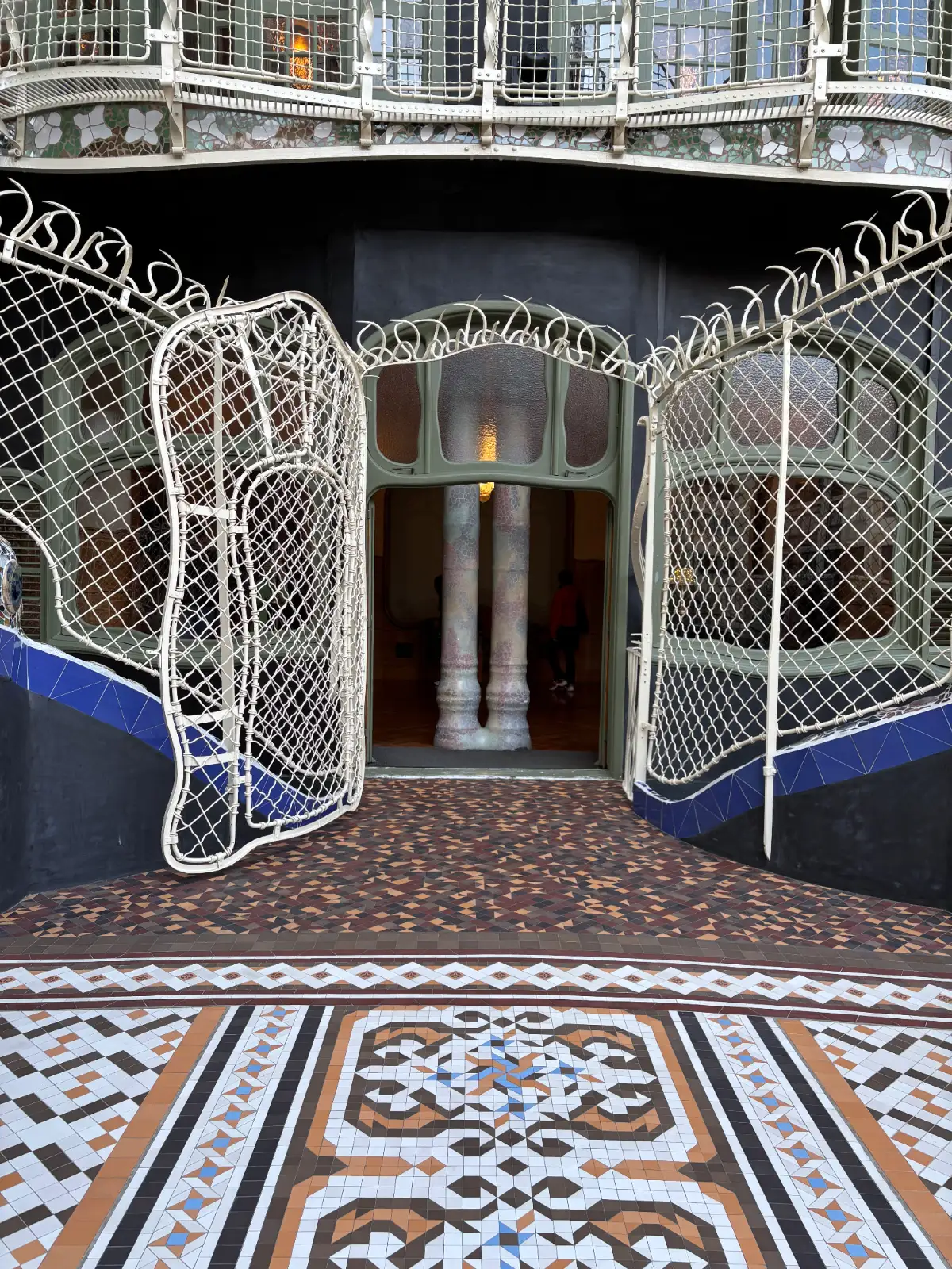 Ornate Art Nouveau entrance of Casa Batlló, with white metal gates, mosaic tile floor, and bone-like columns inside.