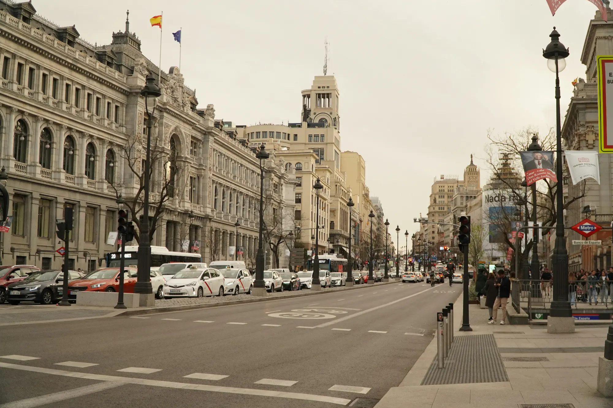 View down the wide Calle de Alcalá in Madrid, lined with grand historic buildings, cars, and people under an overcast sky.