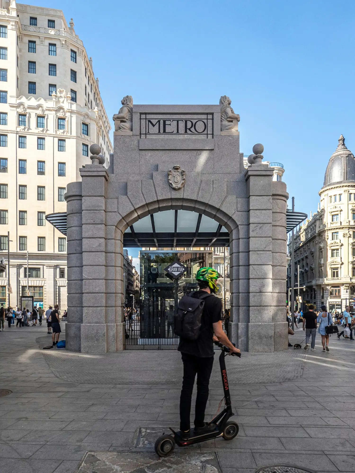 A person on an electric scooter in front of the historic stone entrance to the Gran Vía metro station in Madrid.