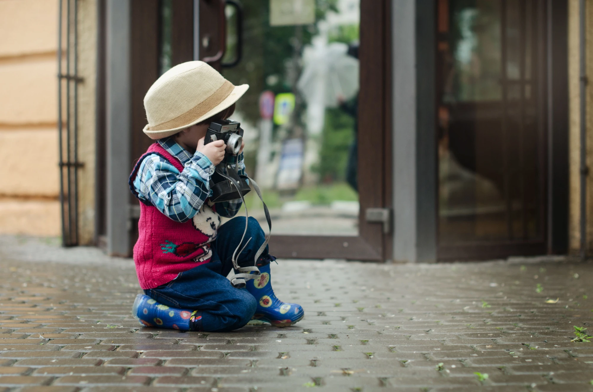 A small child in a hat and rain boots kneels on a wet pavement, holding up an old-fashioned camera.