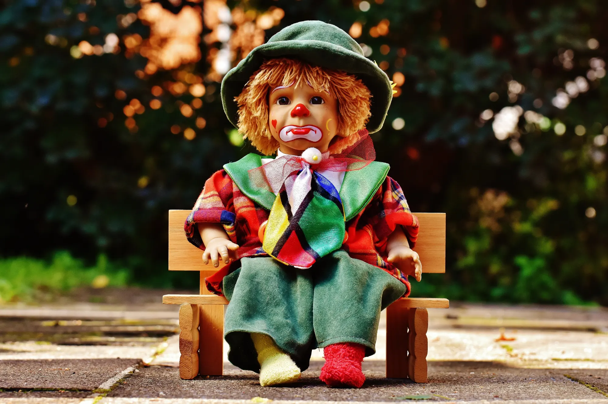 A sad-faced clown doll in a colorful outfit and green hat, sitting on a tiny wooden bench outdoors.
