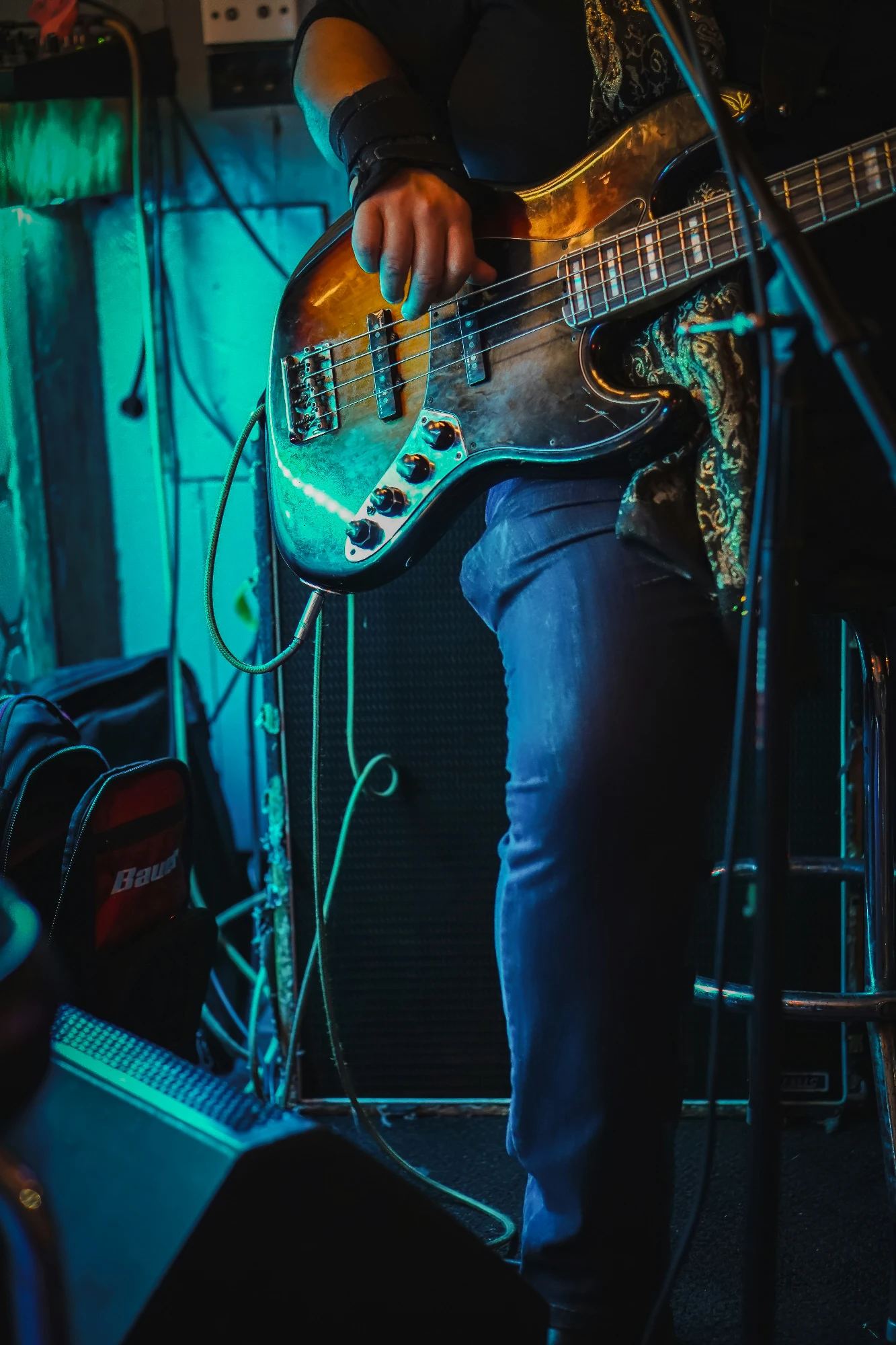 A musician plays a sunburst bass guitar on a dimly lit stage with turquoise lighting.