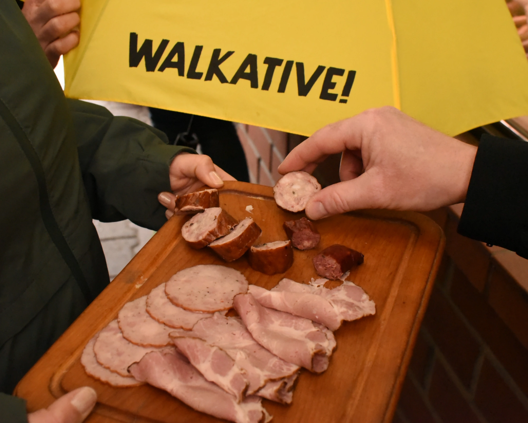 Close-up of people tasting sliced sausage and cold meats from a wooden board under a yellow Walkative umbrella.
