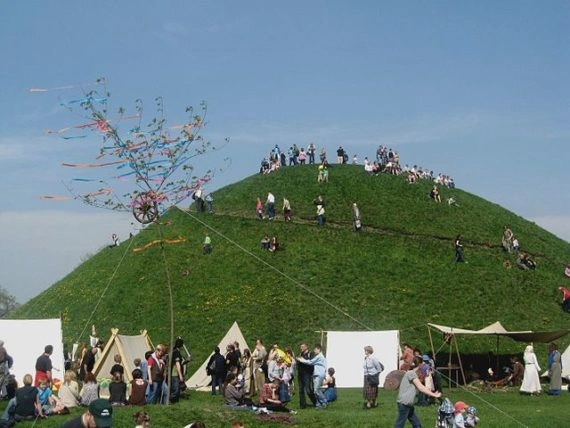 Large green Krakus Mound with many people on its slopes and around tents at the base, under a clear blue sky. A tall pole with colorful ribbons is visible.