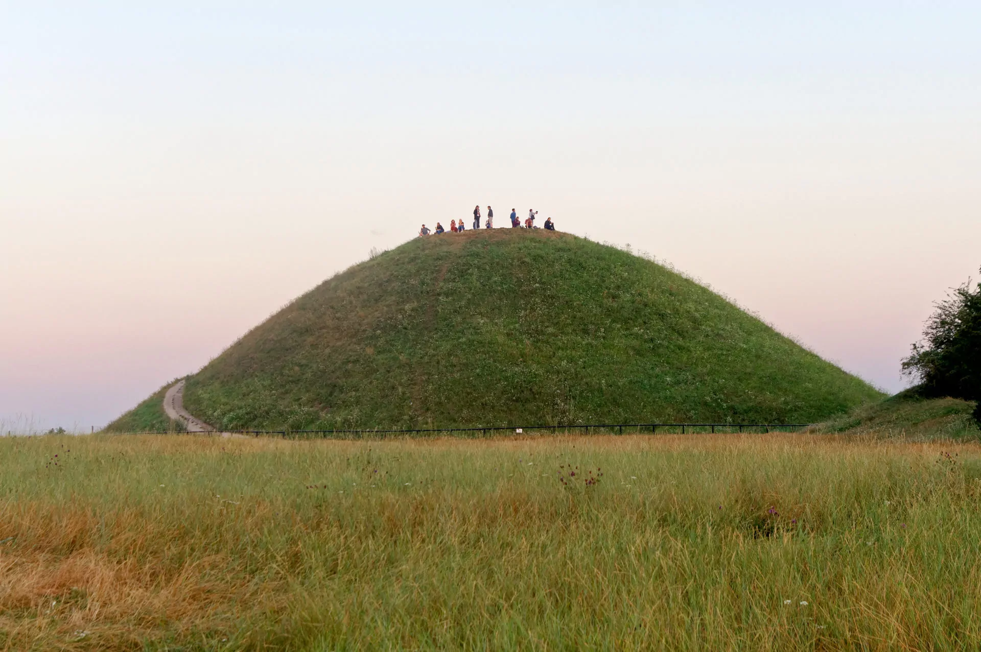 Grassy Krakus Mound in Krakow with a group of people at the summit, a winding path, and dry grass in the foreground under a pale sky.