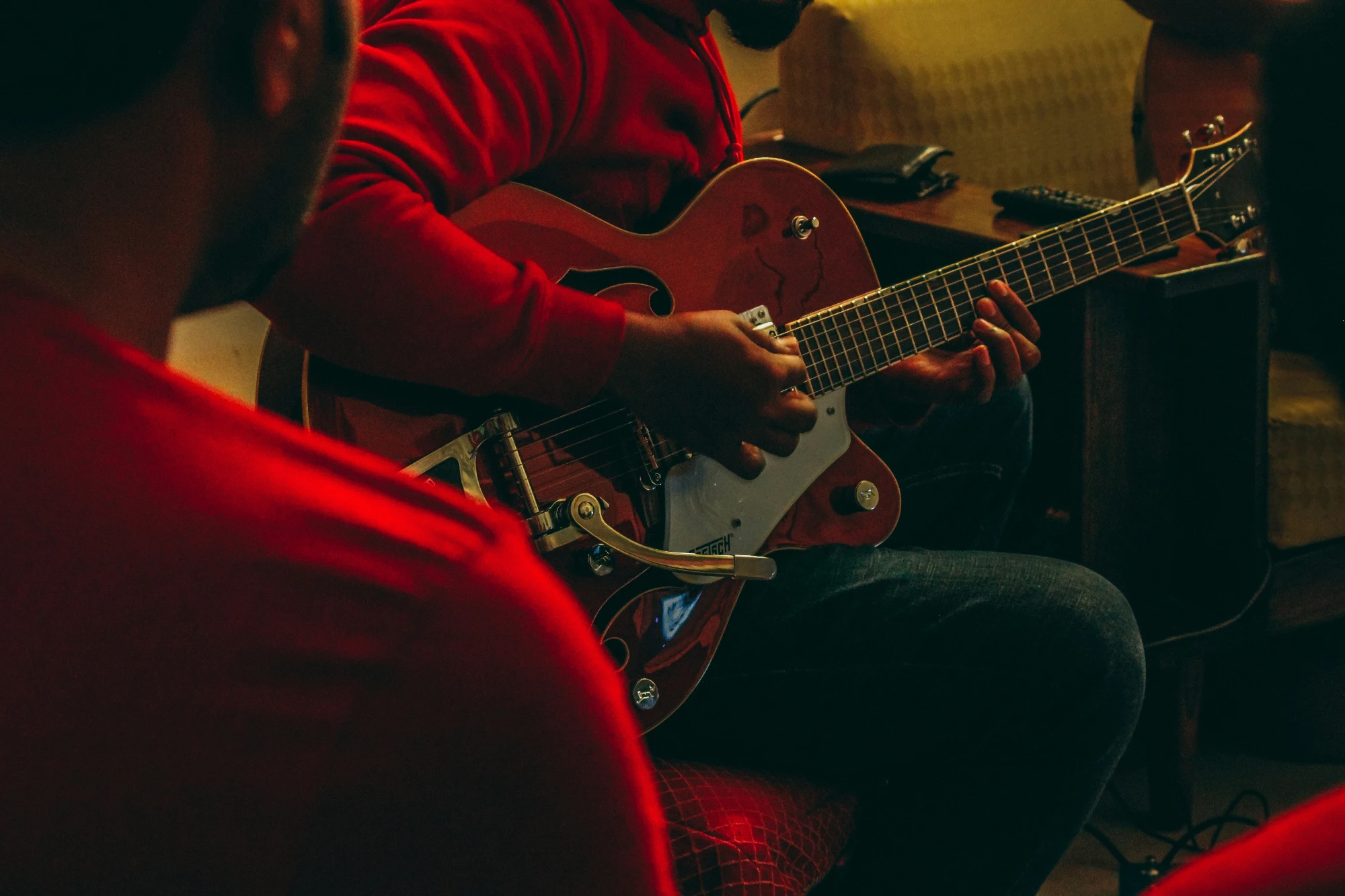 A person in a red hoodie plays a red electric guitar in a dimly lit room, seen over someone's shoulder.