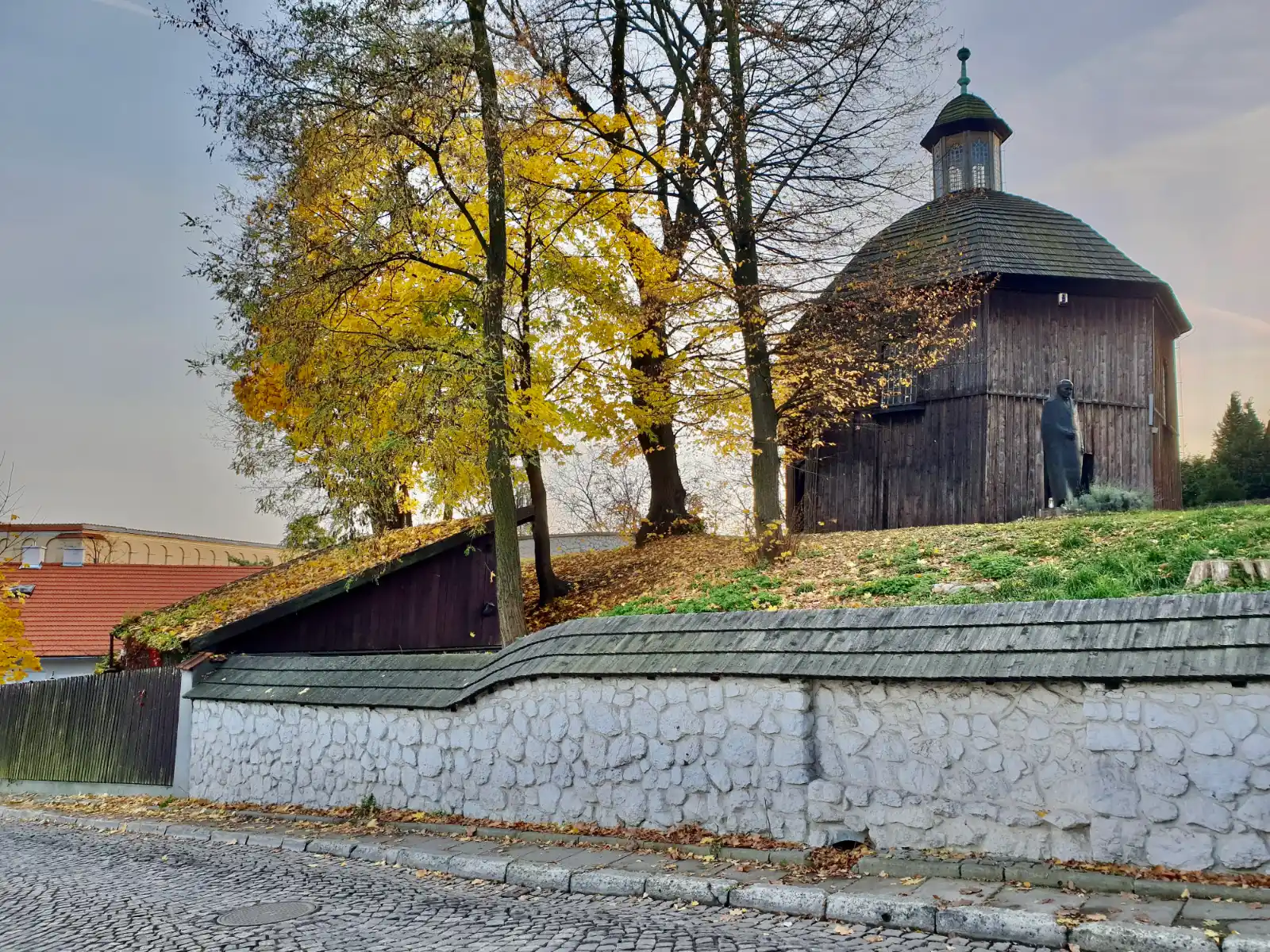An old wooden church on a hill with autumn trees, behind a stone wall on a cobblestone street.