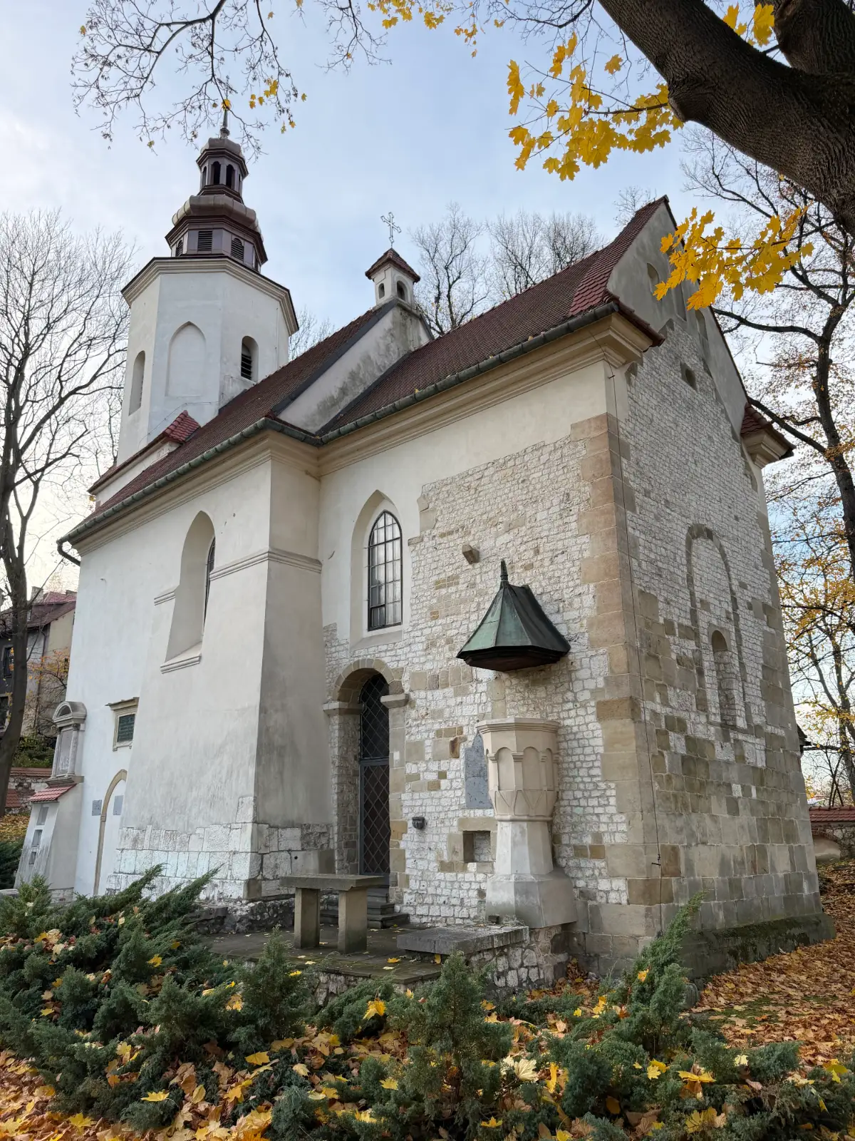A small, white and stone church with a single tower, surrounded by trees with yellow autumn leaves.